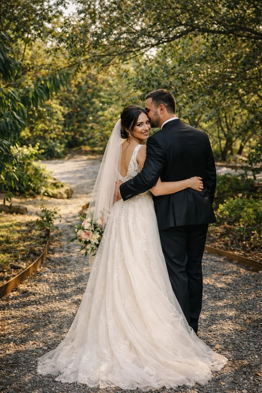Bride and groom in an outdoor setting, embracing and smiling, with the bride holding a bouquet of pink and white roses, surrounded by trees and greenery.