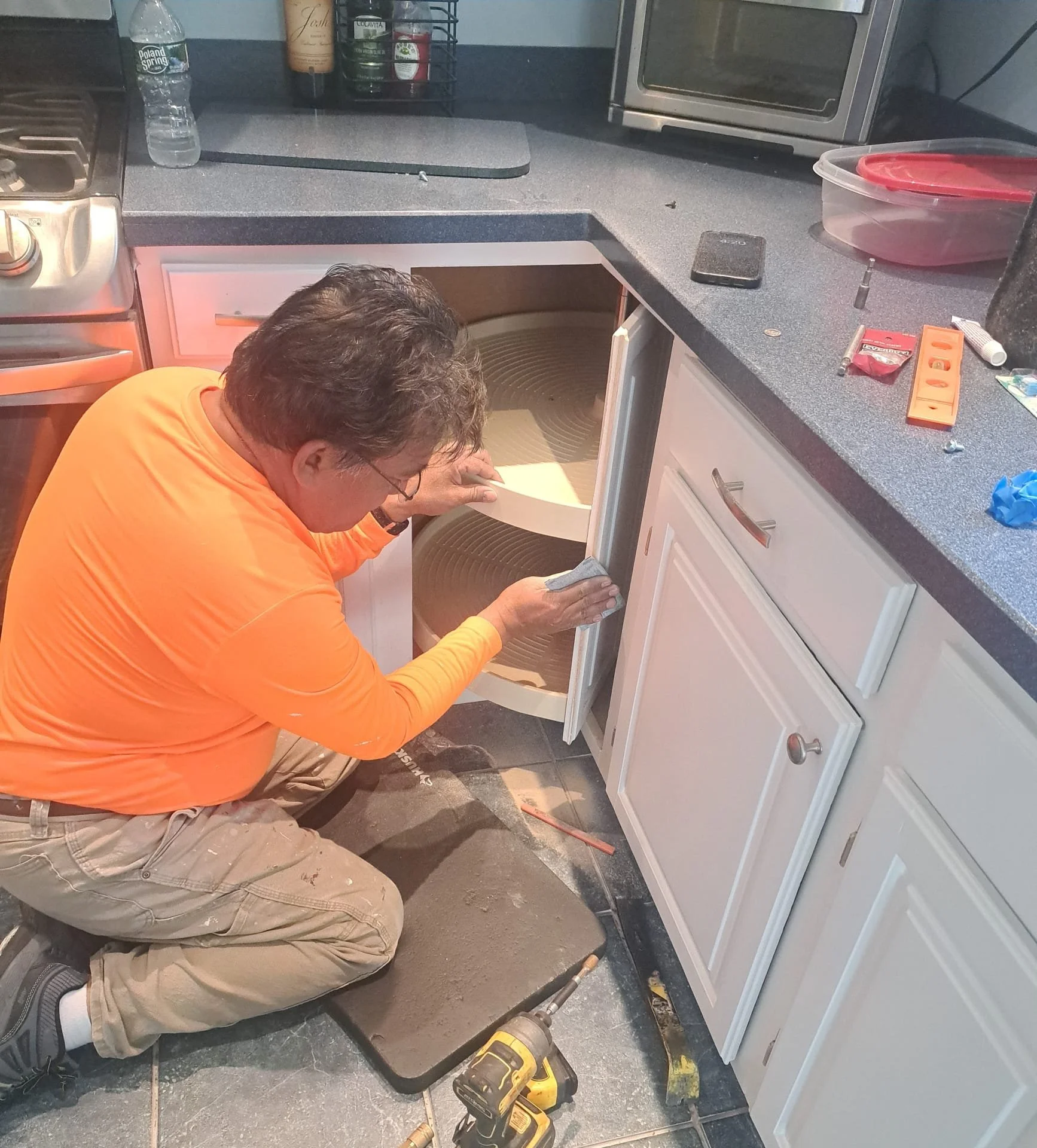 A man in an orange shirt and beige pants installing or repairing kitchen cabinets. He is kneeling on a floor mat, working with tools, with the cabinet door open, revealing the interior of the cabinet. The kitchen counter has various tools and items on it, including a level, a tube of adhesive, and a drill. There is a water bottle, some condiments, and an oven in the background.
