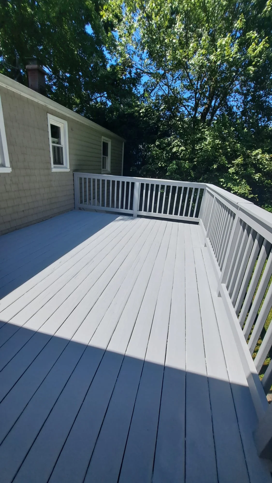 A wooden outdoor deck with white painted flooring and white railing, attached to a house with beige siding and windows, surrounded by green trees under a partly cloudy blue sky.