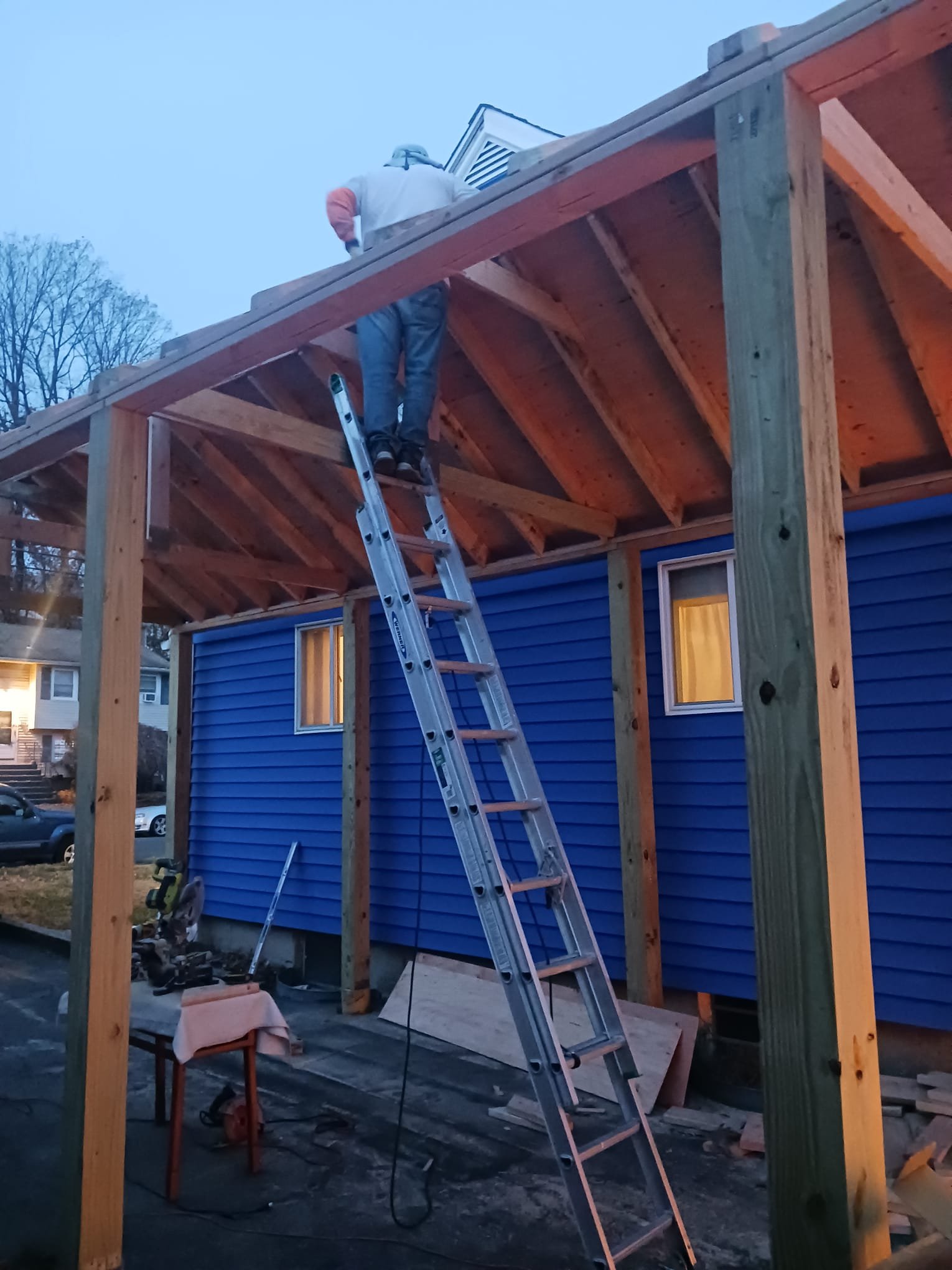 A person working on the roof of a house under construction, standing on a ladder, with wooden beams and a blue house in the background.