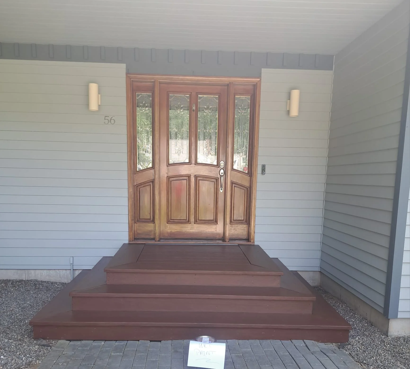 Front porch with wooden steps leading to a wooden door with glass panels and stained glass details, with light gray siding walls and wall-mounted lights on either side of the door.