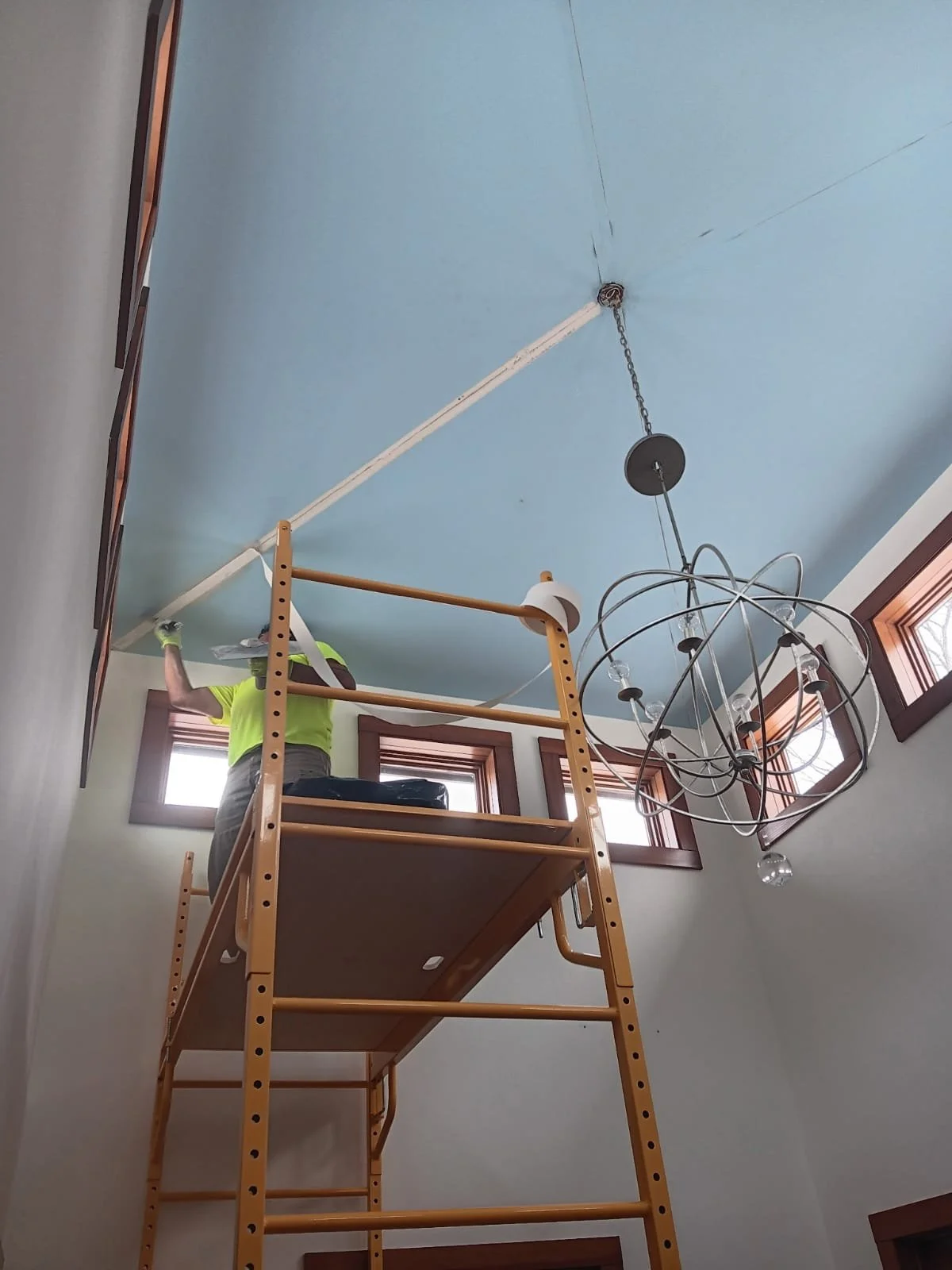 Person on a ladder working on the ceiling, with a chandelier hanging and windows with wooden trim behind.