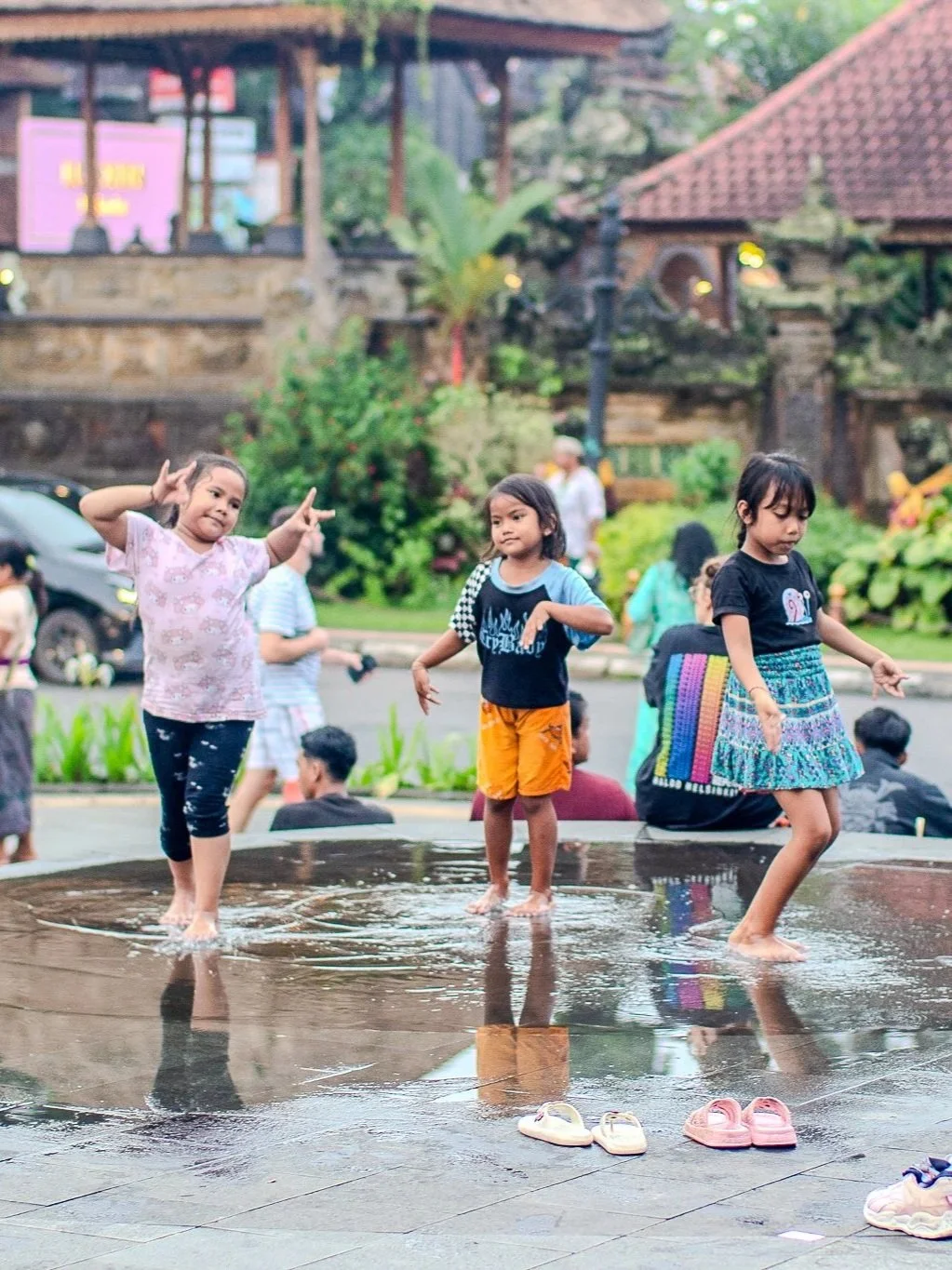 Three children playing and dancing on a rainy, shallow water-covered surface outdoors, with shoes off in front of them. There's greenery and buildings in the background.