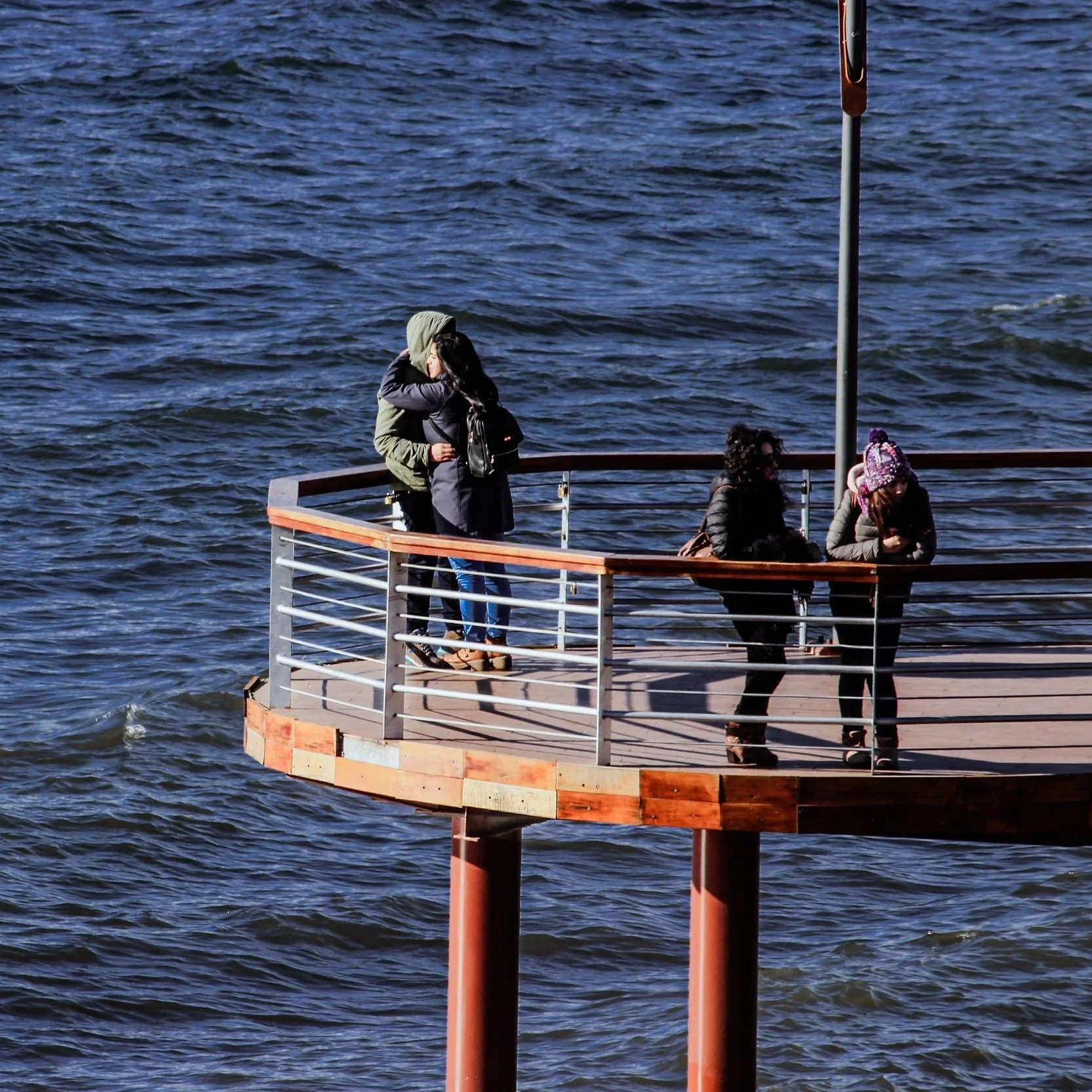 Four people standing and sitting on a wooden platform overlooking a body of water, some of them wearing warm clothing and hats
