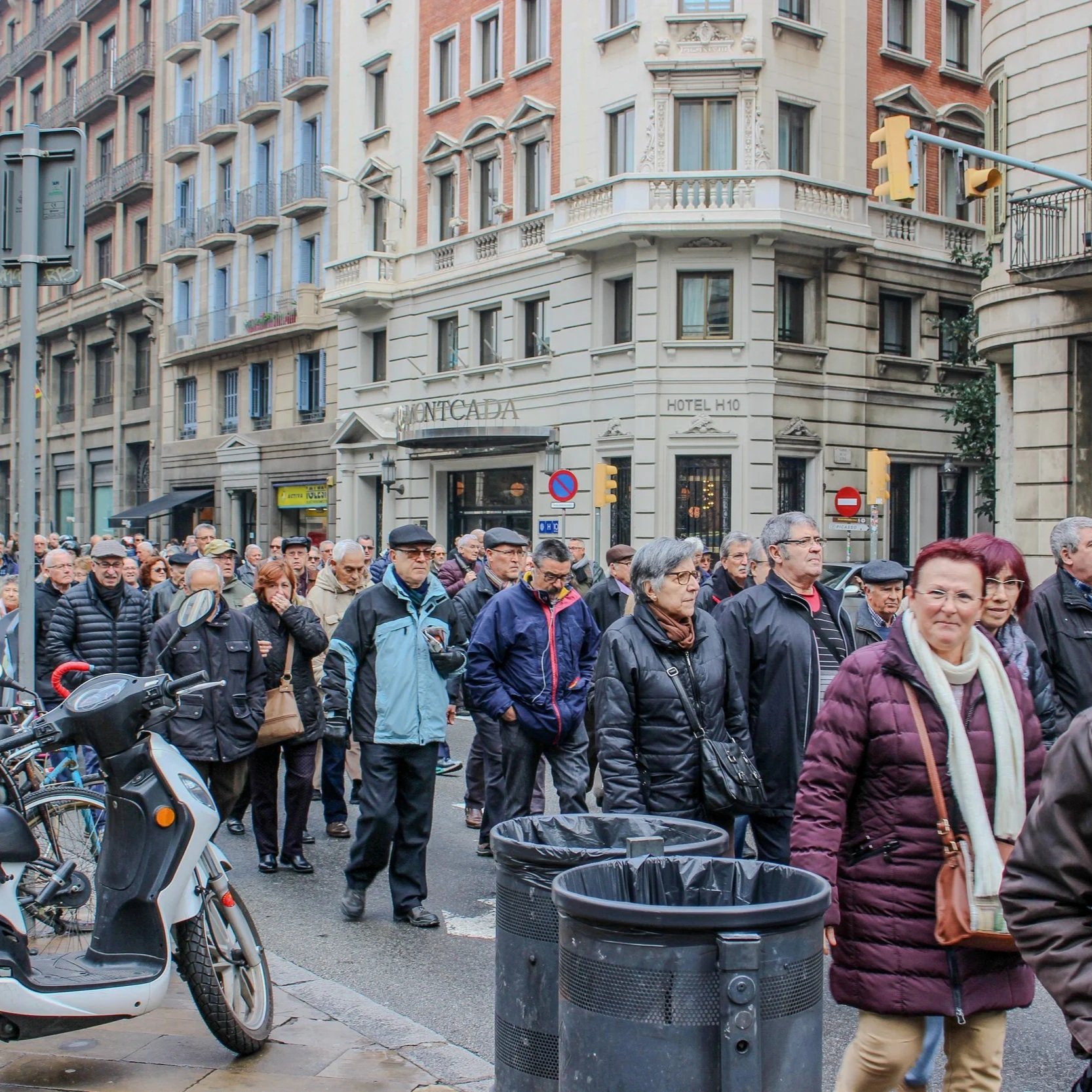 A group of people walking on a city street in front of a building with ornate architecture. There are bicycles and a scooter parked nearby, and trash cans are visible in the foreground. The scene suggests a busy urban area, possibly during an event o