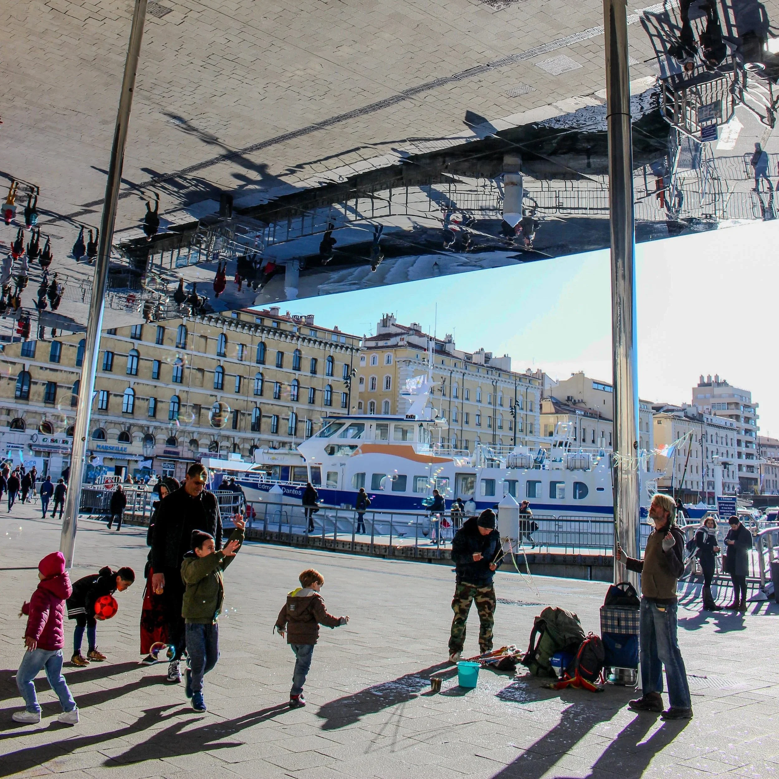 People walking and standing on a city street near a docked boat, with shadows cast on the sidewalk from nearby structures and street posts.