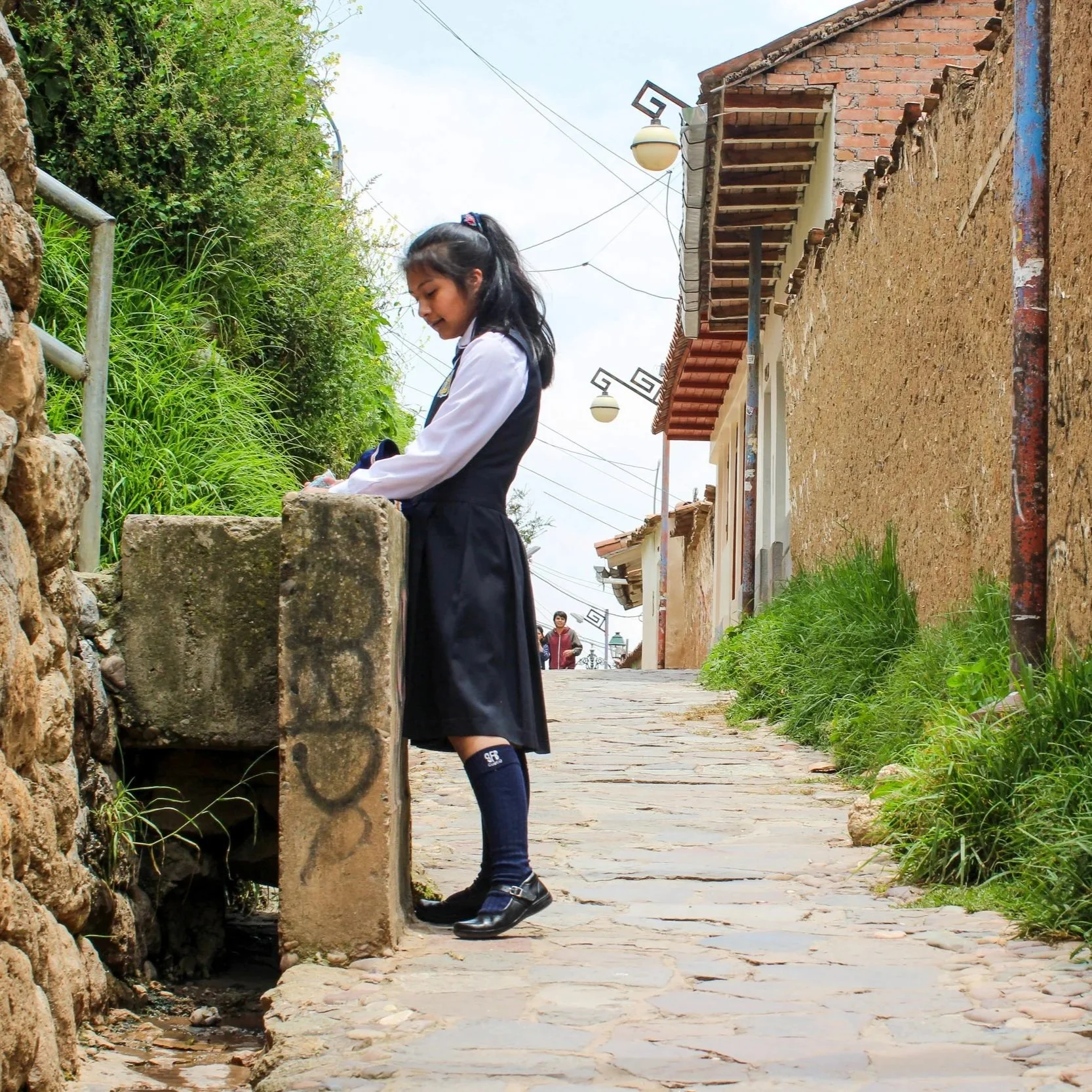 A young girl in a school uniform, including a navy skirt, white shirt, and navy socks, is standing on a cobblestone sidewalk and looking down, near a stone wall with green foliage on the other side. There are houses with brick and stucco walls, tiled
