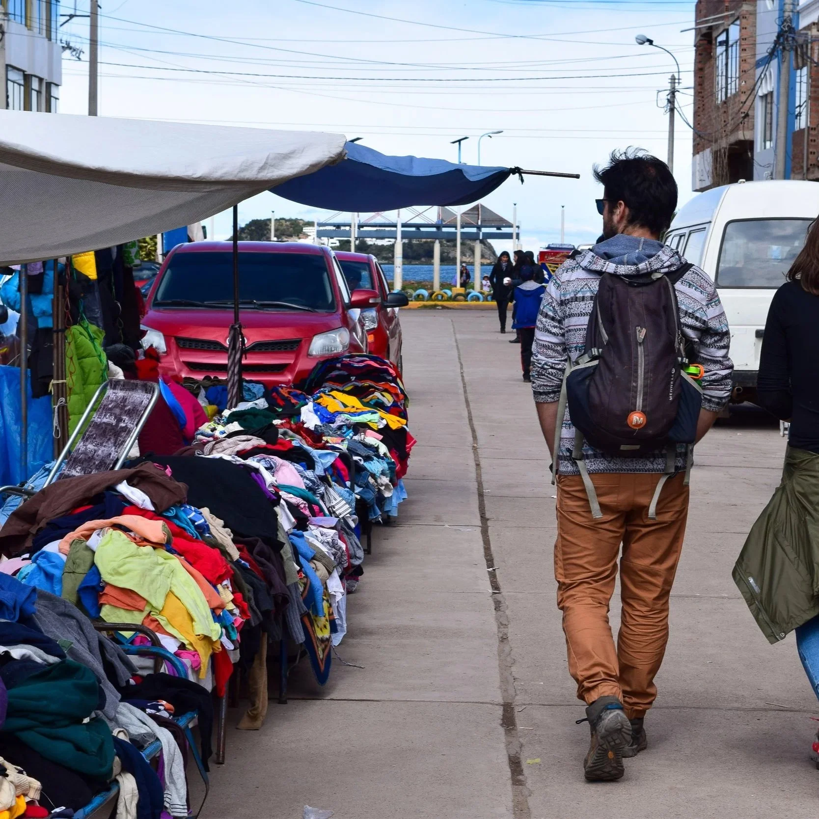 Street market scene with clothing and gear for sale on tables, a person walking away with a backpack, and a waterfront with a bridge in the background.