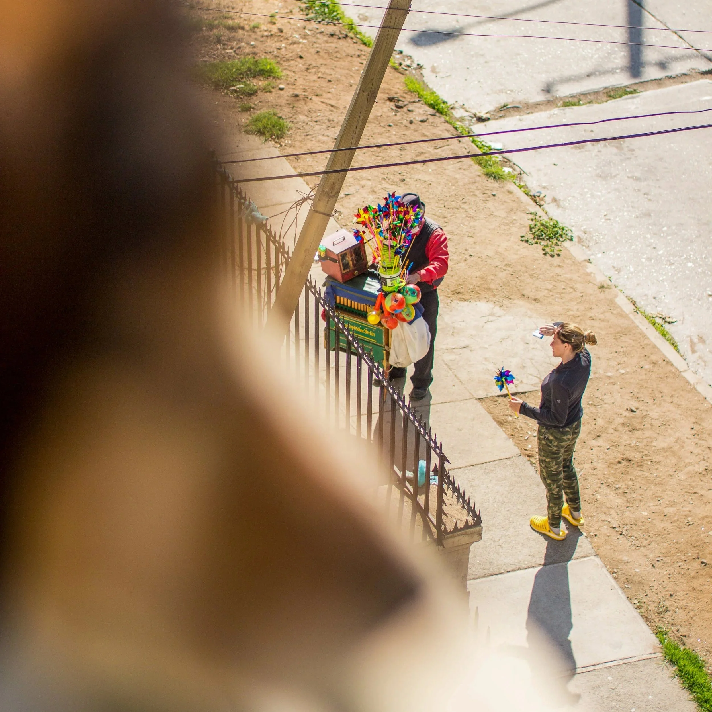A person holding a container filled with colorful pinwheels on a sidewalk next to a metal fence, with another person nearby, and a woman behind them taking a photo. The photo is partially obscured by a finger in the foreground.