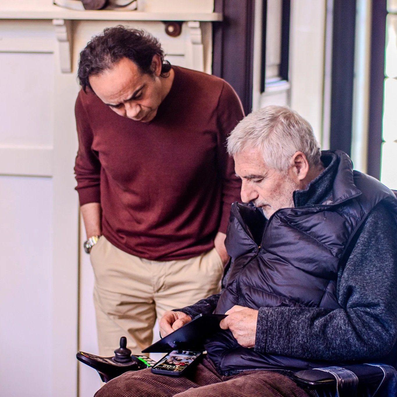 Two men, one standing and one seated, looking at a phone together in a cozy indoor setting.