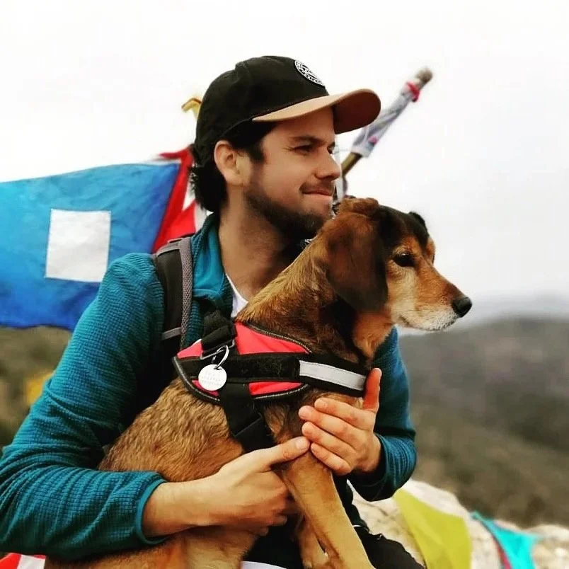 A man holding a brown and black dog outdoors with a partially visible colorful flag in the background.