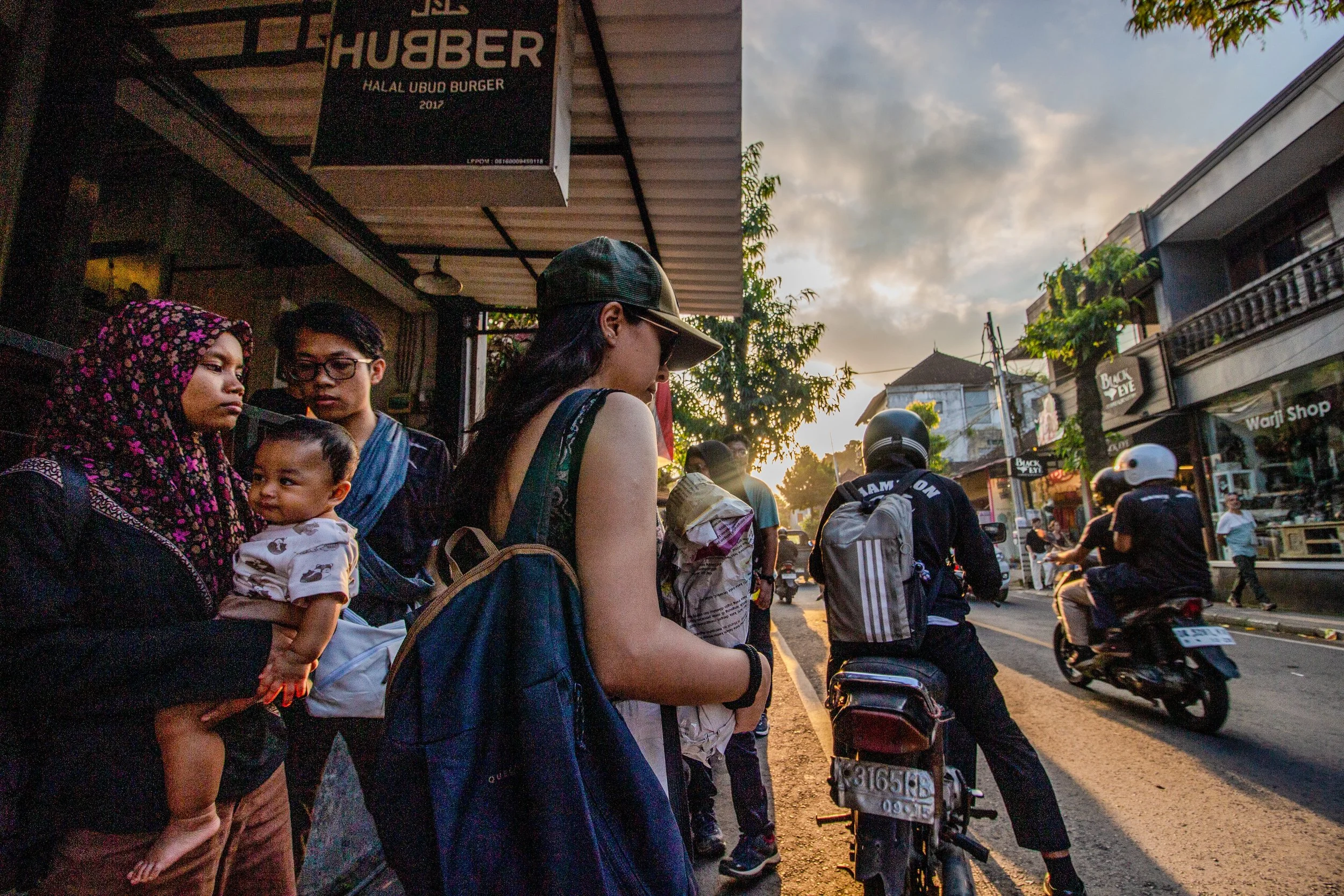 People waiting at a bus stop during sunset on a busy street, with motorcycles passing by and commercial buildings in the background.