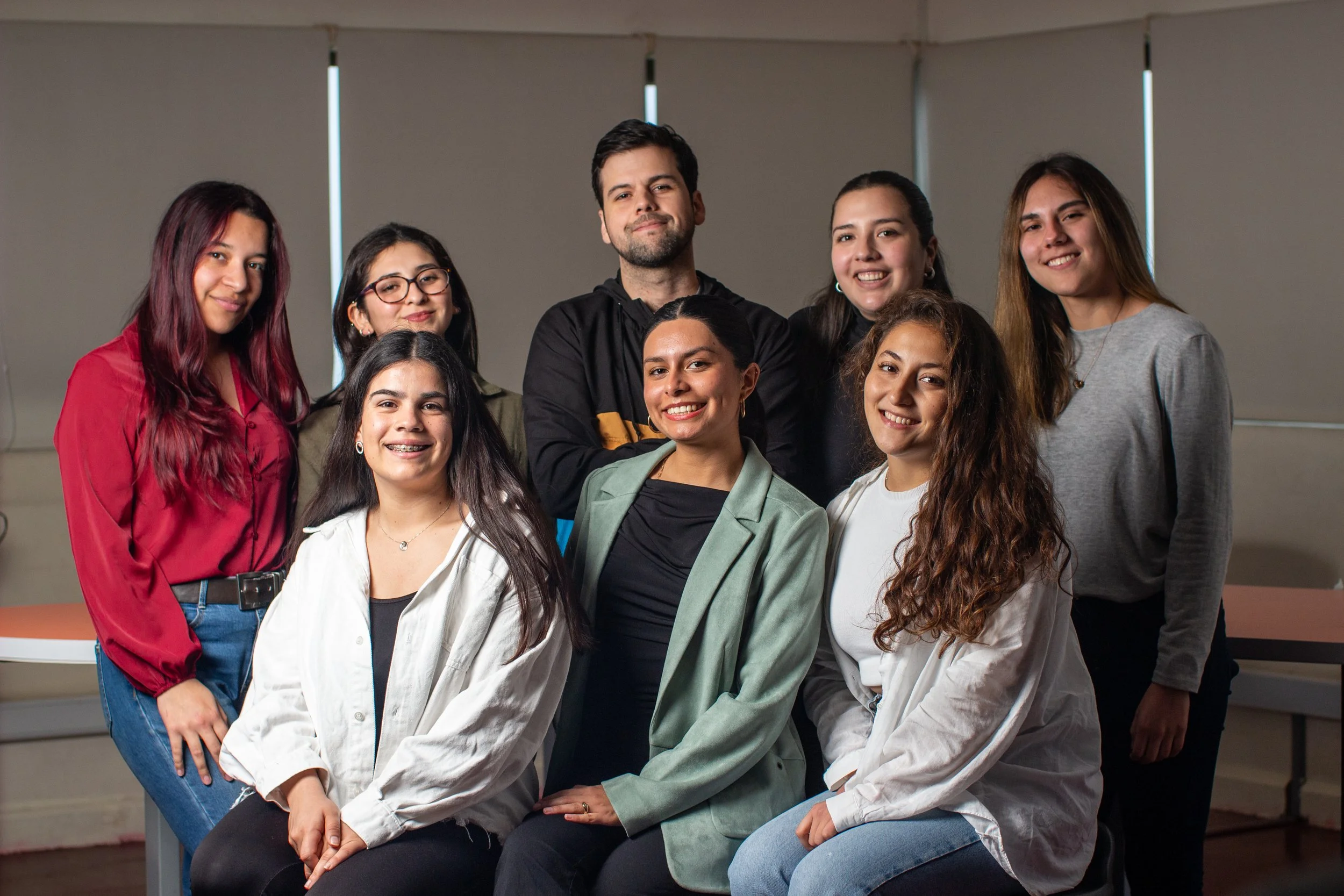 Group of eight young adults posing together in an indoor setting, some sitting and some standing, smiling at the camera.