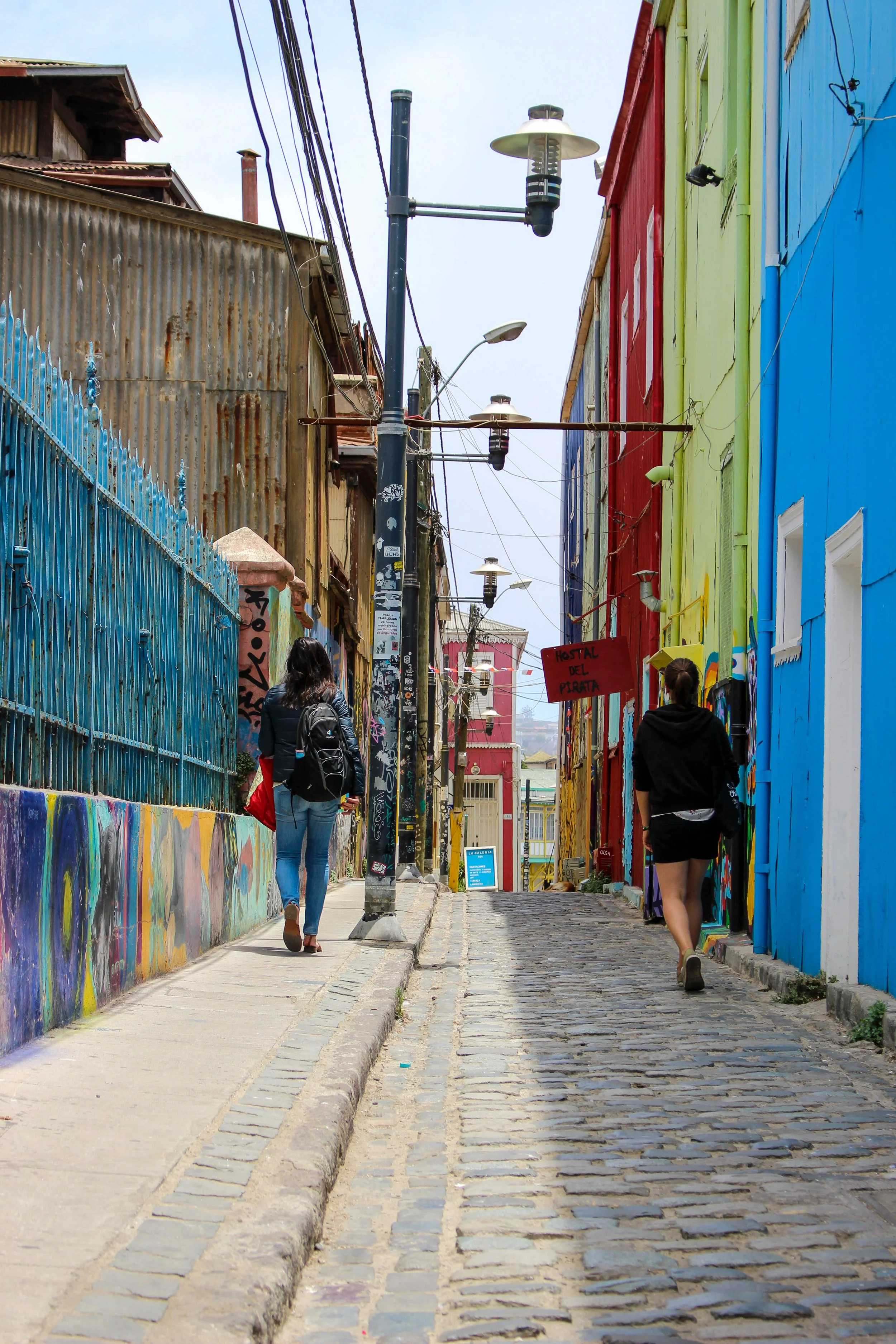 A colorful street with buildings painted in bright blue, red, green, and yellow, with graffiti on the walls. There are two women walking on the cobblestone street, one with a backpack and the other in shorts. Ernes streetlights are overhead, and the sky is cloudy.