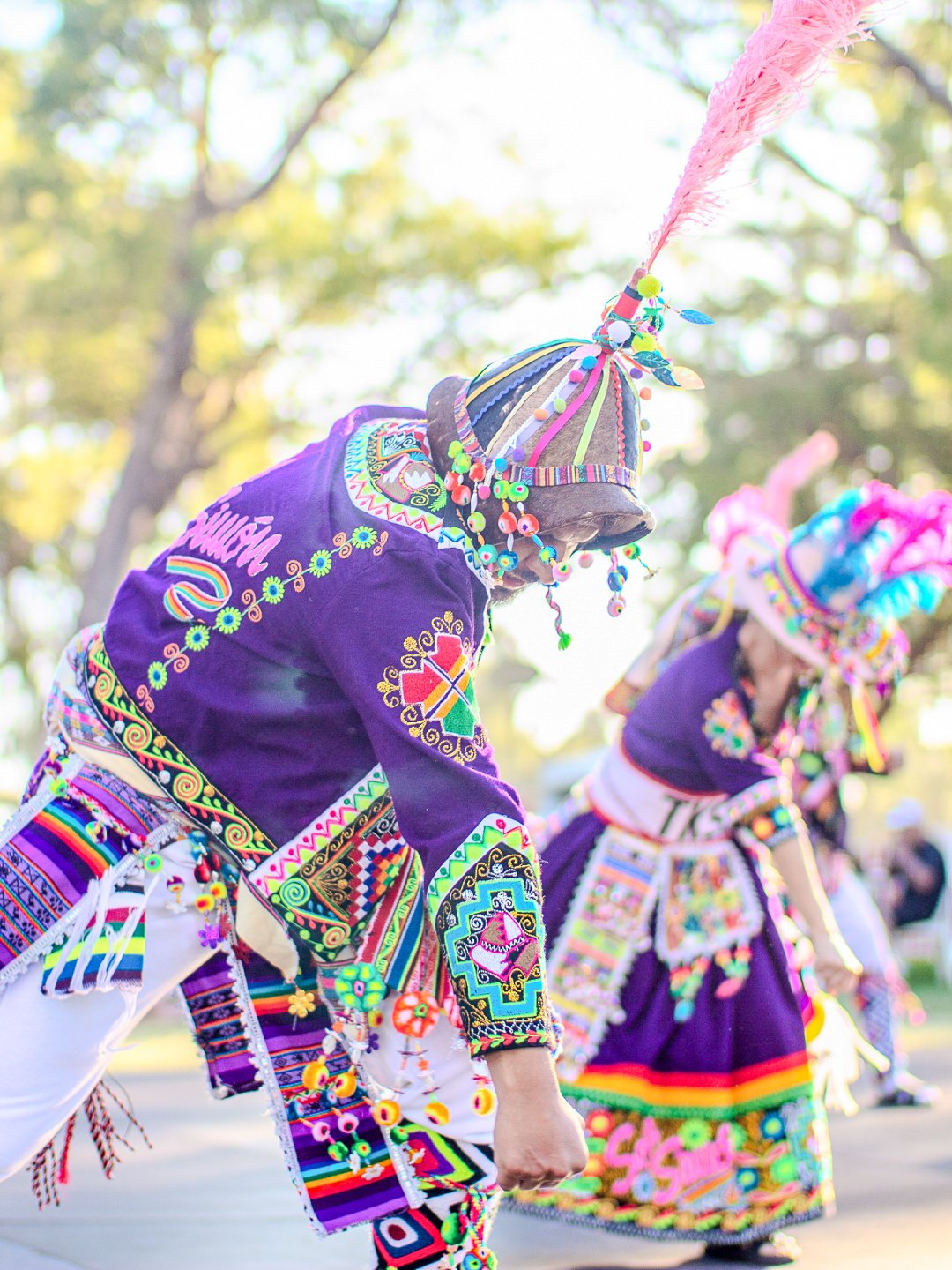 Two people dressed in colorful, traditional, embroidered clothing performing a dance outdoors, with trees in the background.