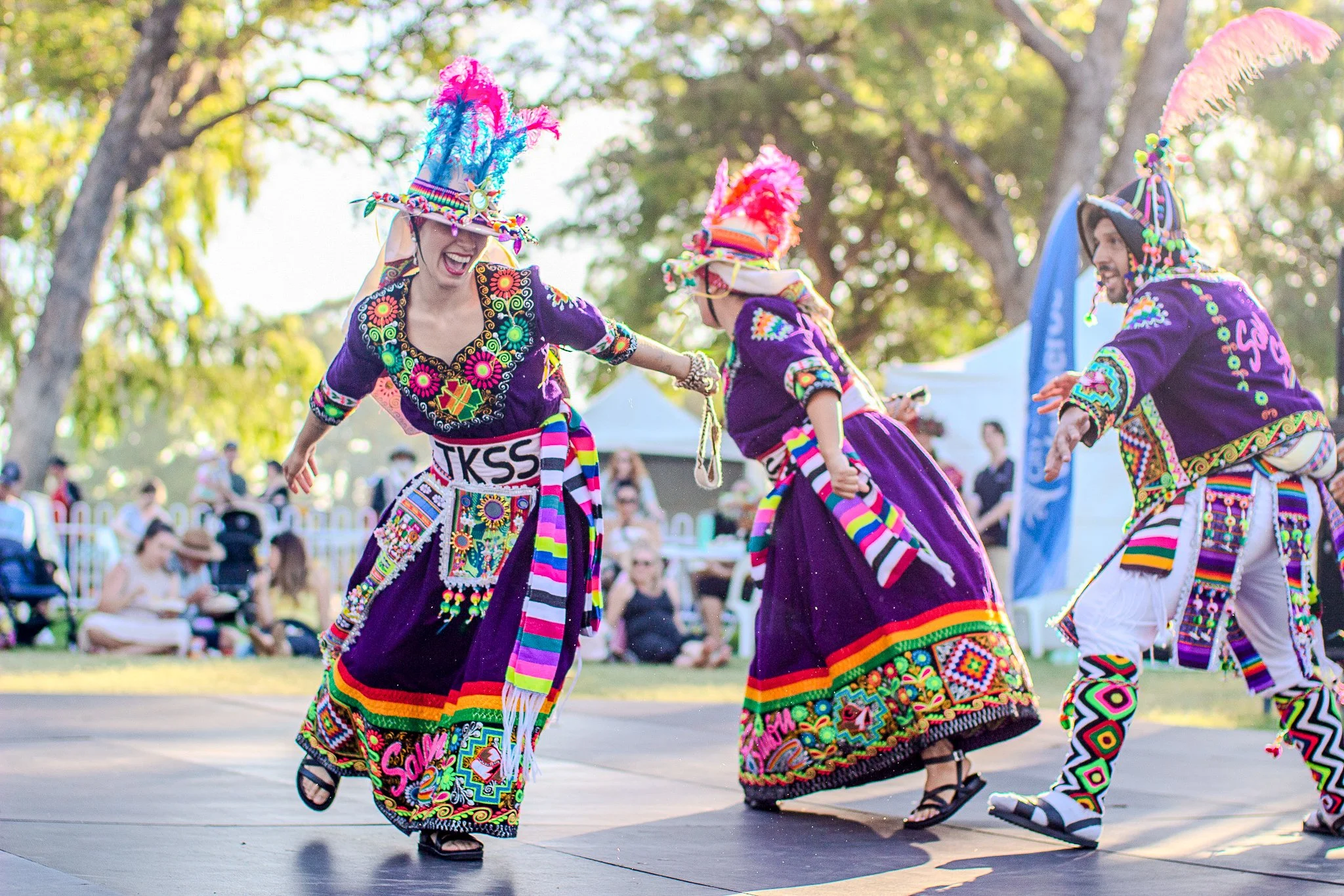 People dressed in bright, colorful traditional outfits, performing a lively dance outdoors during the daytime.