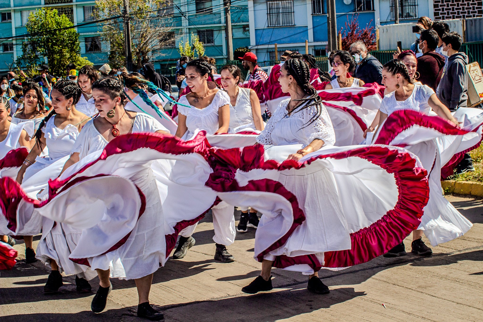 A group of women in white outfits dancing with large red and white skirts during a parade on a sunny day in an urban area.