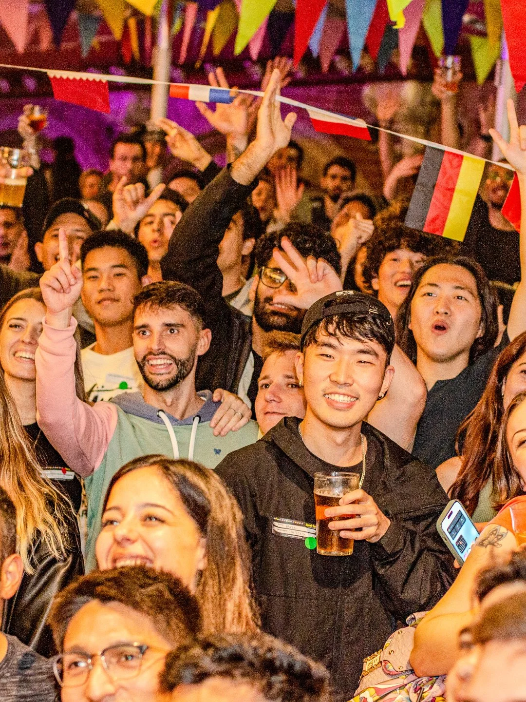 Crowd of diverse young adults at a lively party or celebration, with flags and colorful decorations overhead, some smiling and holding drinks, others making gestures and enjoying the event.