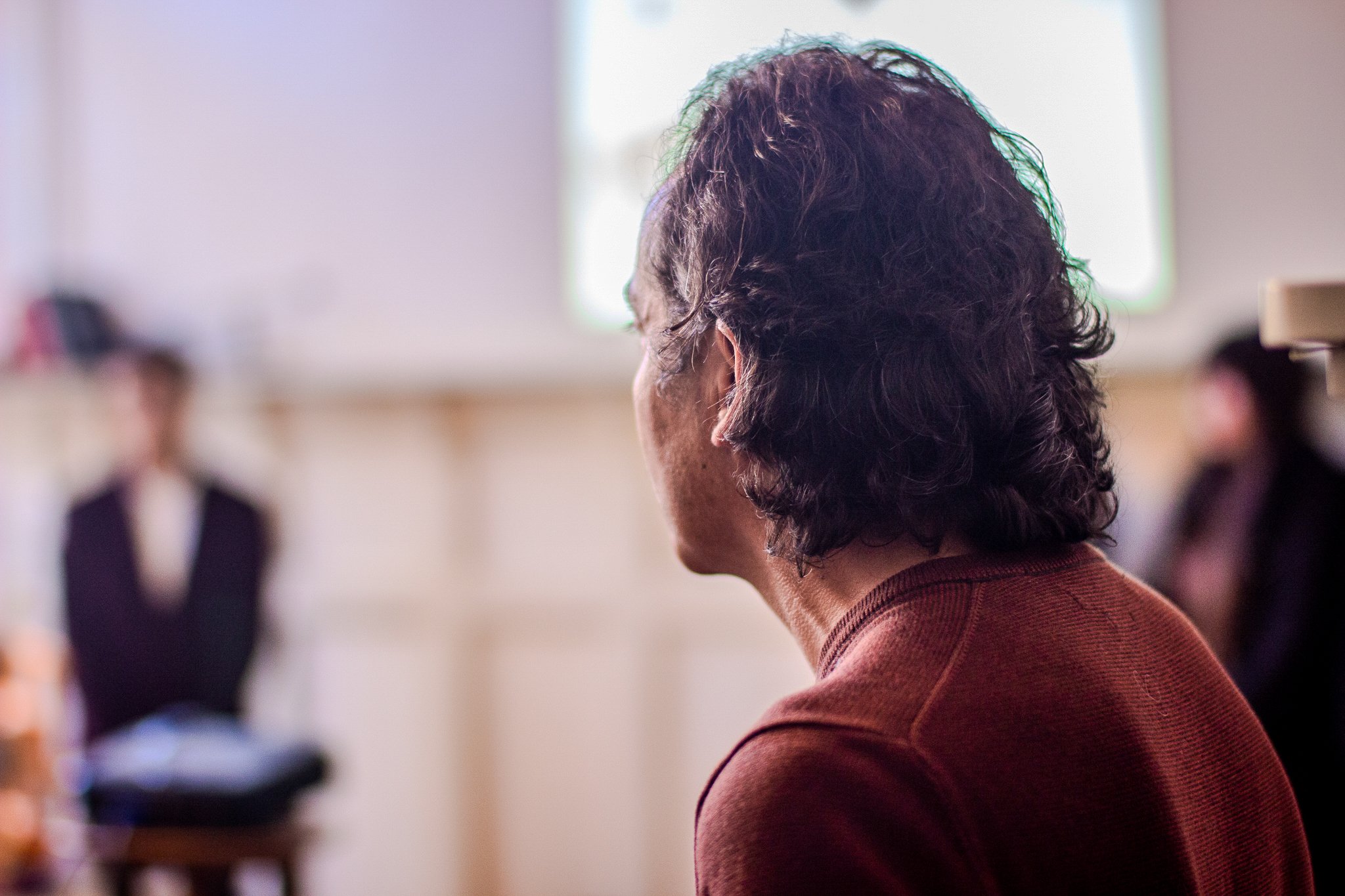 A person with curly dark hair, wearing a maroon sweater, seen from behind, in a room with blurred figures of other people.