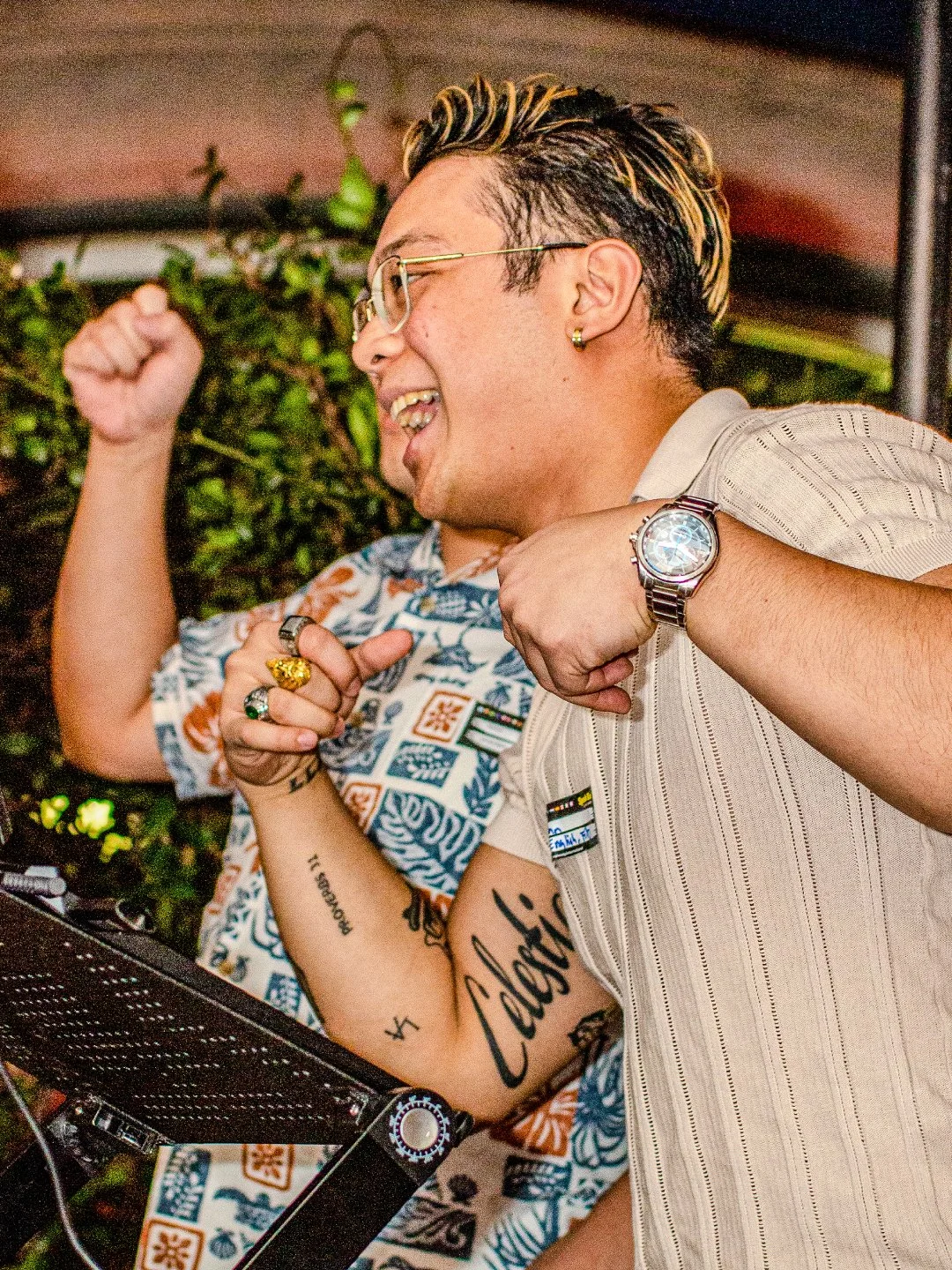 Two people celebrating and smiling joyfully, with one flexing his arm and the other clenching his fist, near DJ equipment.
