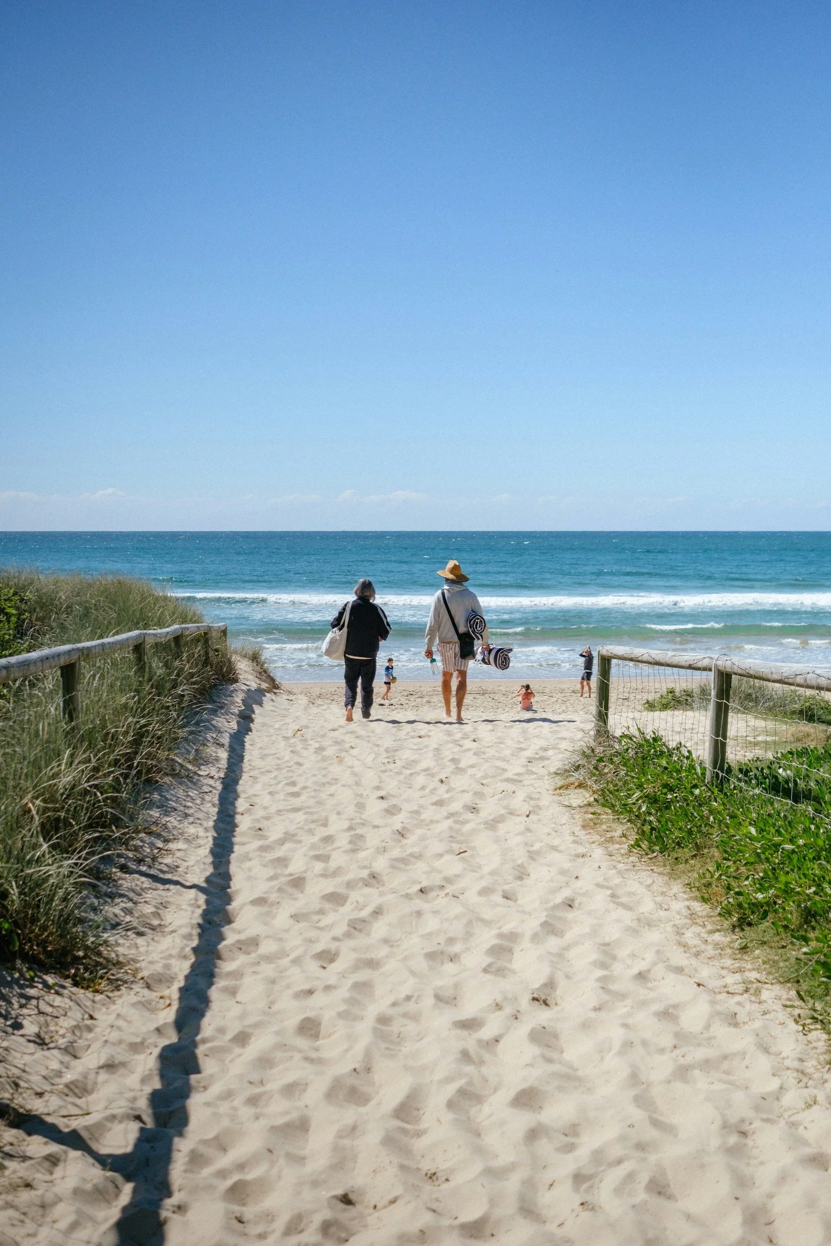 Two people walking on a sandy beach path towards the ocean, with a grassy area on the left and a barrier on the right under a clear blue sky.