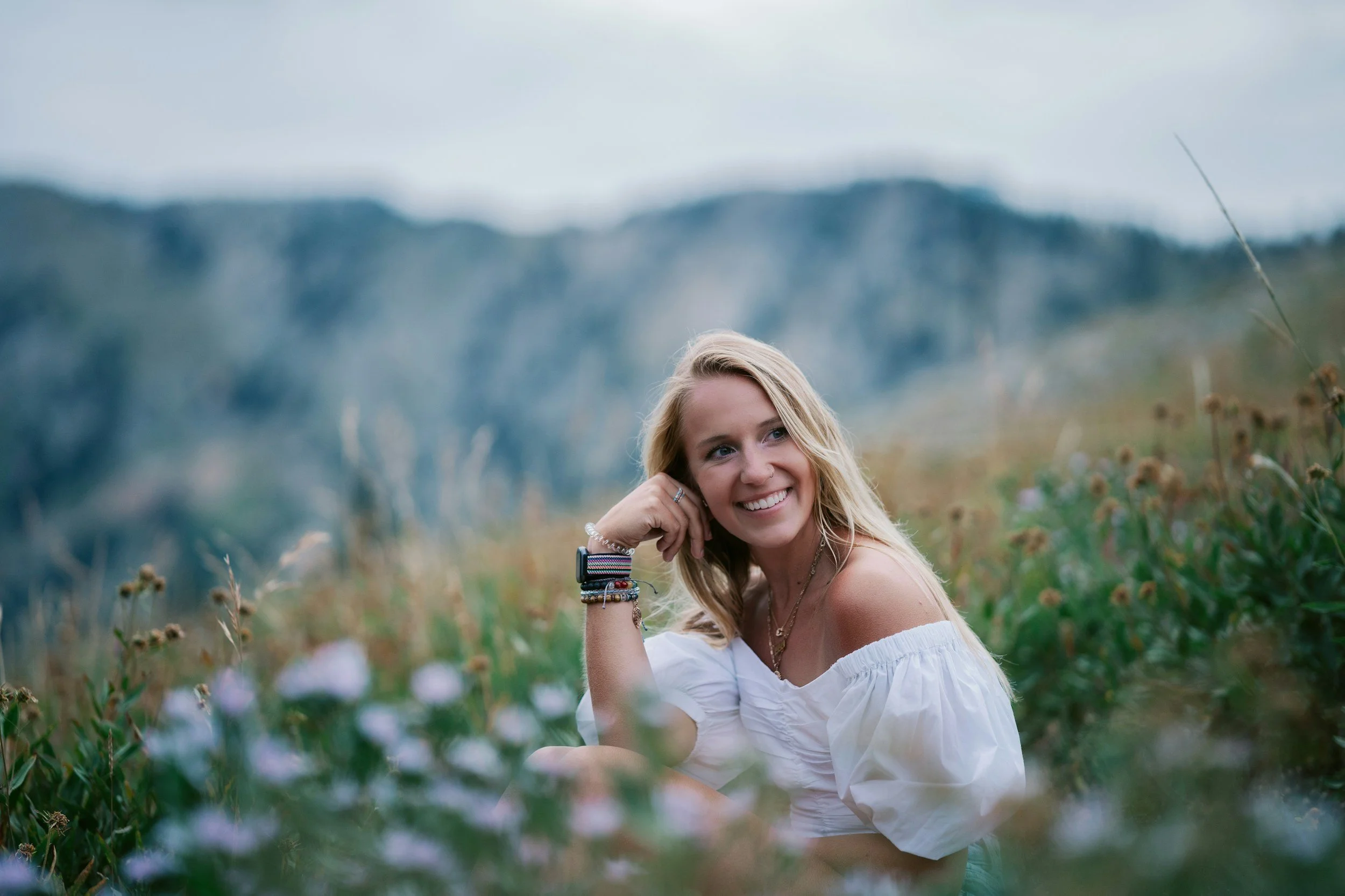 A young woman with blonde hair, smiling, sitting in a field of flowers with mountains in the background, wearing a white off-shoulder top and colorful bracelets.