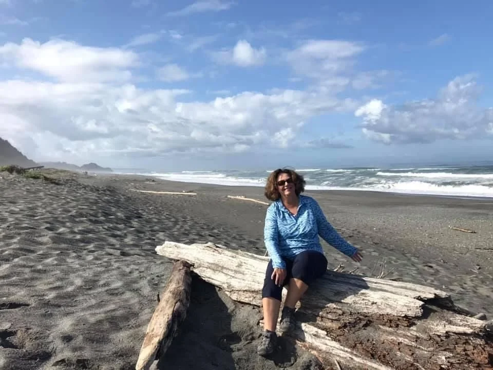 A woman sitting on a large piece of driftwood on a sandy beach with ocean waves and a partly cloudy sky in the background.
