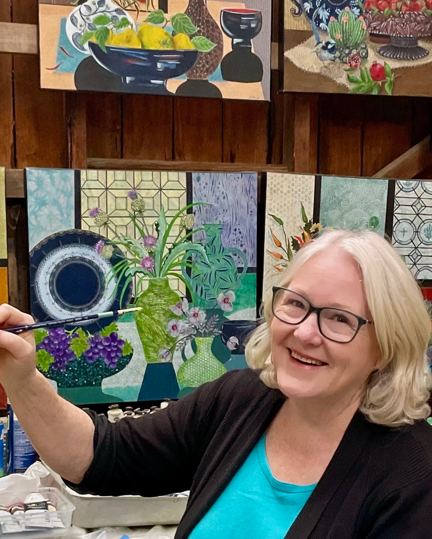 A woman with glasses smiling while holding a pen and working on a colorful acrylic painting of vases, flowers, and plates in an art studio.
