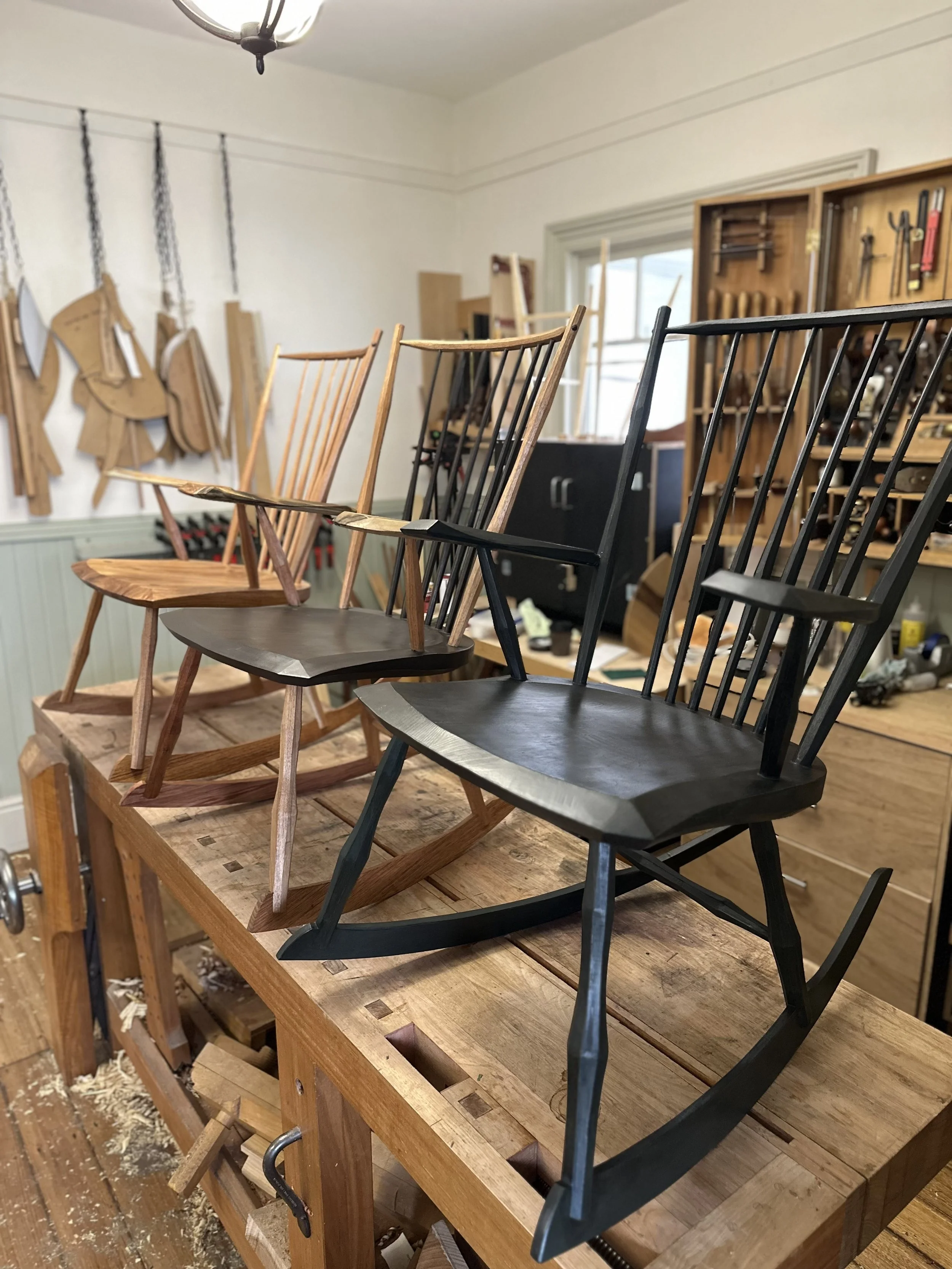 A woodworking workshop with various chairs in different stages of being assembled, sitting on a sturdy wooden workbench. The background includes woodworking tools and equipment on wall-mounted shelves and pegboards.