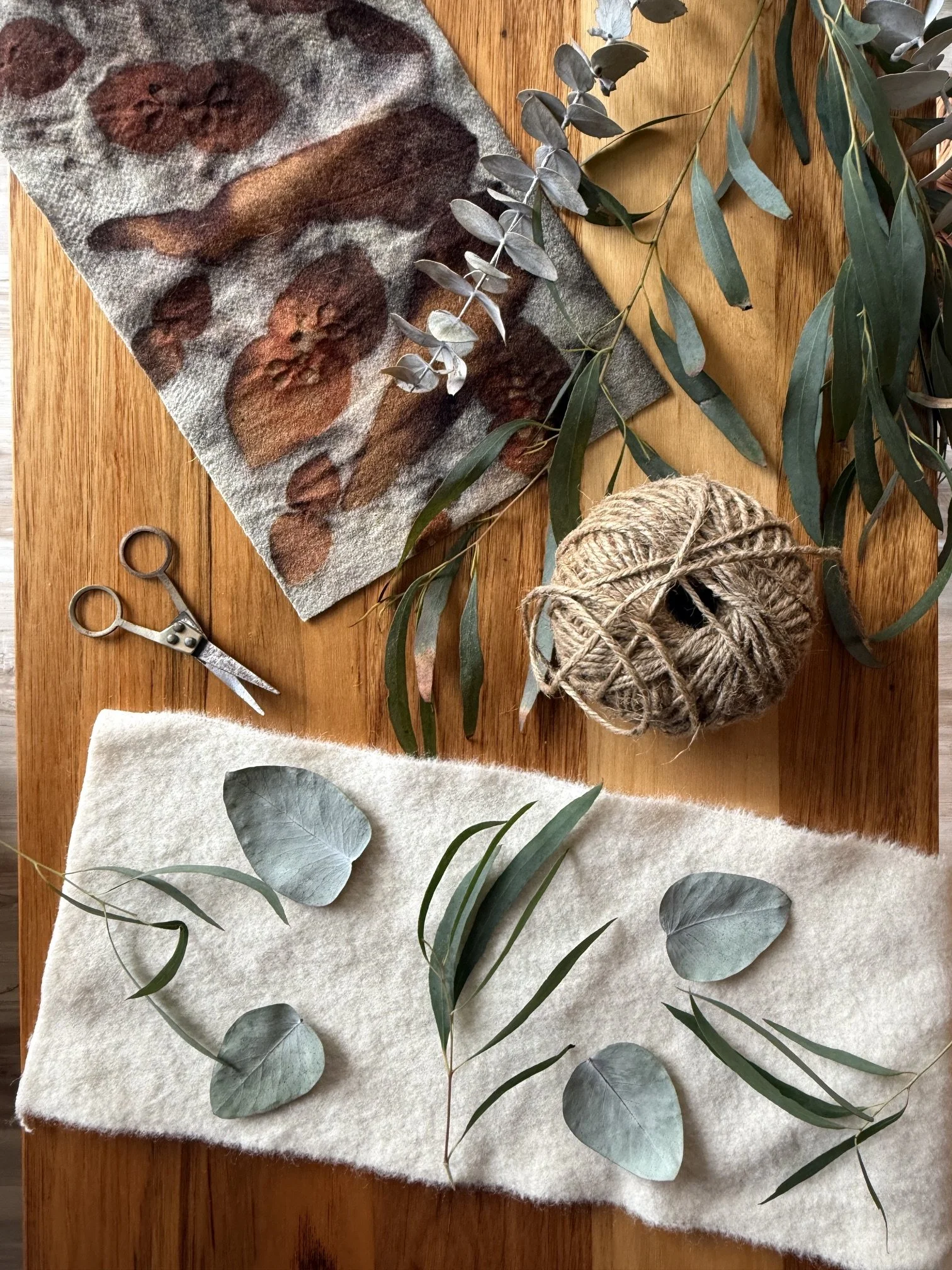 Eucalyptus branches on a table with scissors, a ball of twine, and felt with painted leaves and circles.