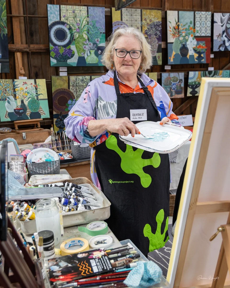 An elderly woman with glasses, wearing a colorful shirt and black apron with green paint splash design, standing in an art studio. She is holding a palette with light blue watercolor paint, surrounded by painting supplies and completed floral artworks displayed on the wooden walls behind her.