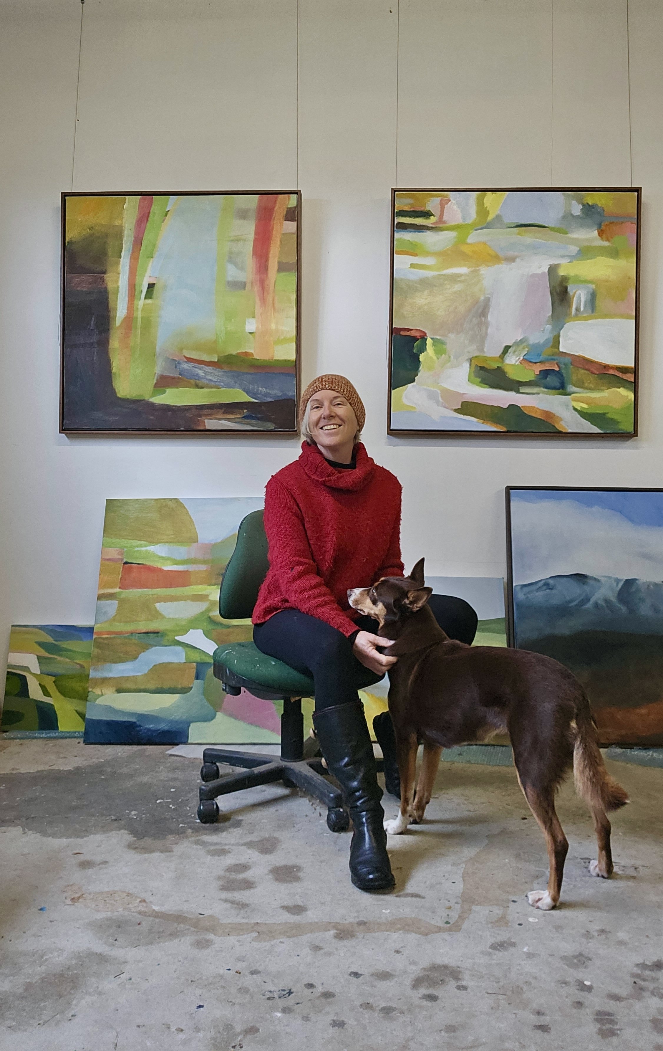 A woman in a red sweater and black boots sitting on a green chair, smiling, with a brown dog standing in front of her, inside an art studio. Behind them, colorful abstract paintings are displayed on the wall and leaning against the wall.