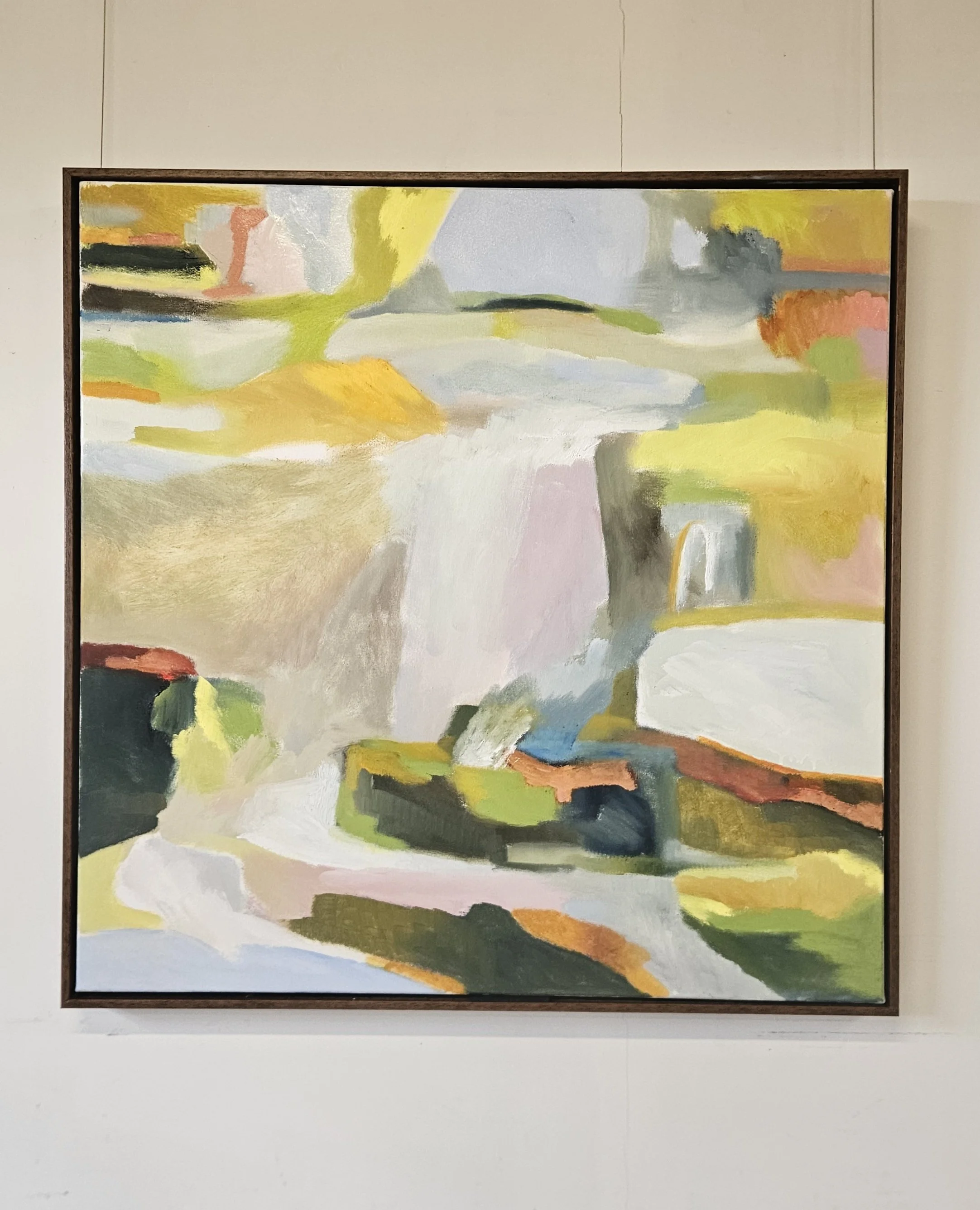 Abstract landscape painting with pastel colors, including yellow, green, gray, black, white, and touches of orange, framed and hung on a plain white wall.