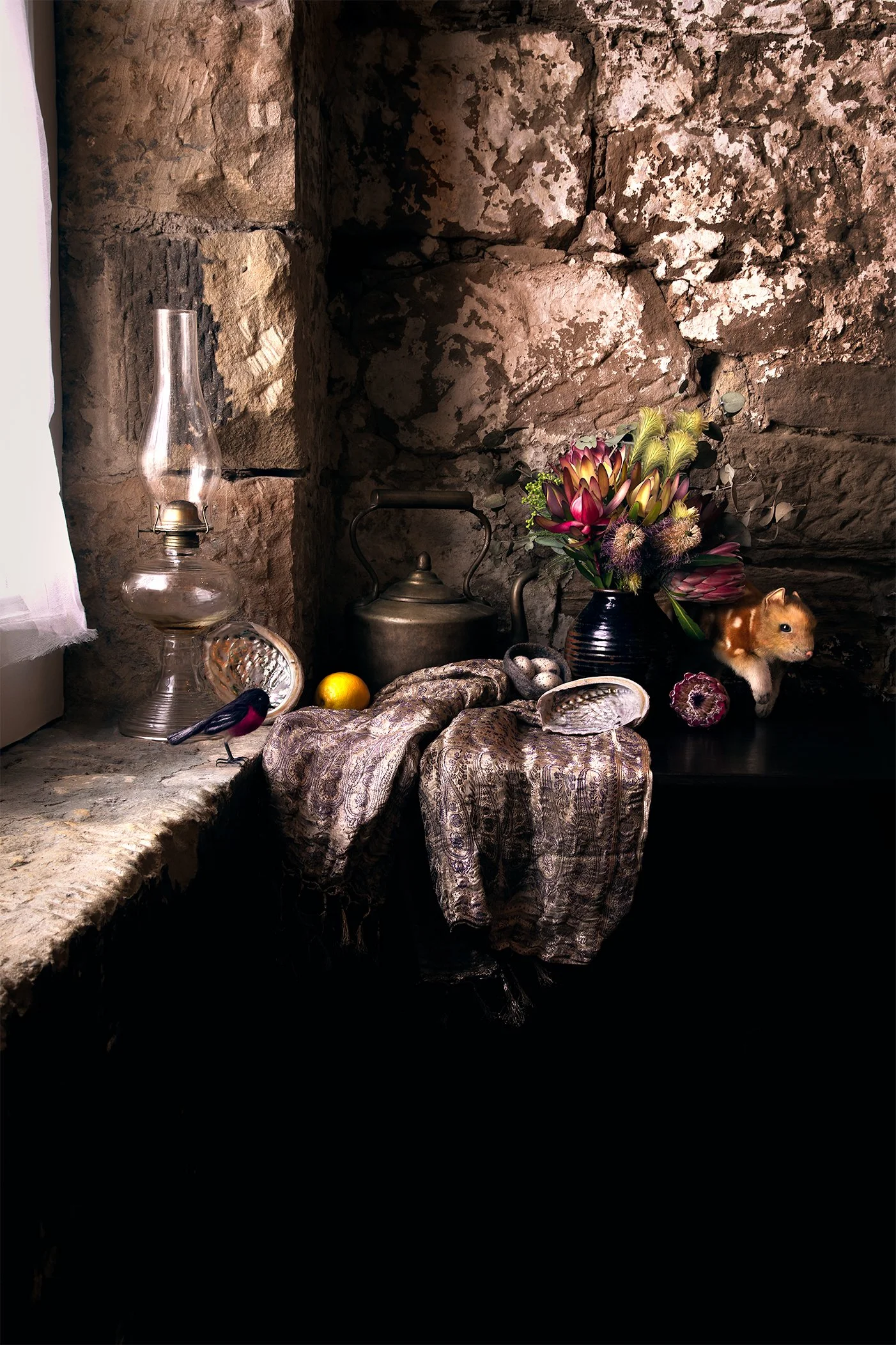 A rustic table with a paisley-patterned cloth, a vintage oil lantern, a brass teapot, a black vase with colorful flowers, a small guinea pig, a yellow lemon, seashells, and dried flowers, all set against a stone wall background.