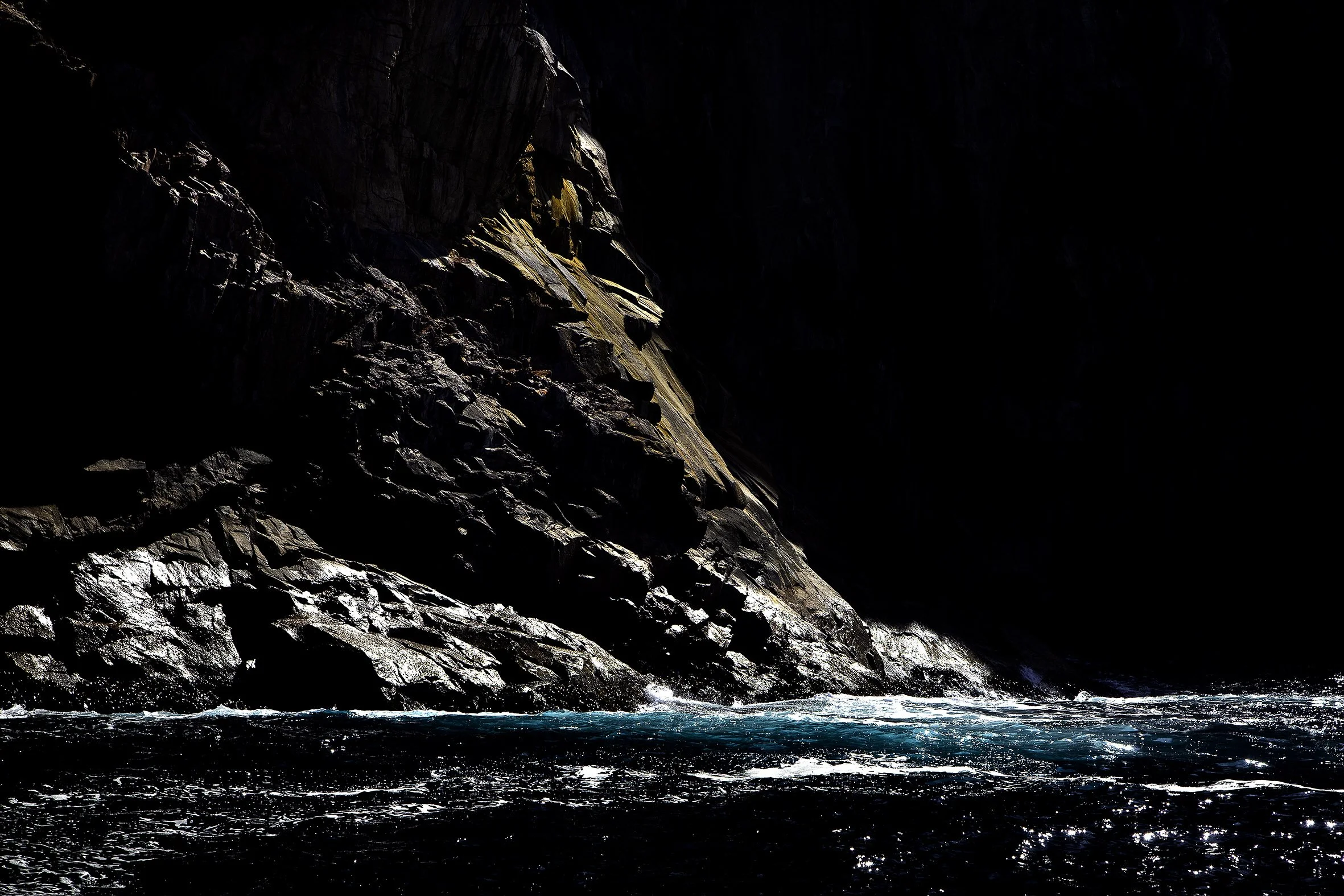 Dark rocky cliffs with some sunlight hitting them above of turbulent blue water.
