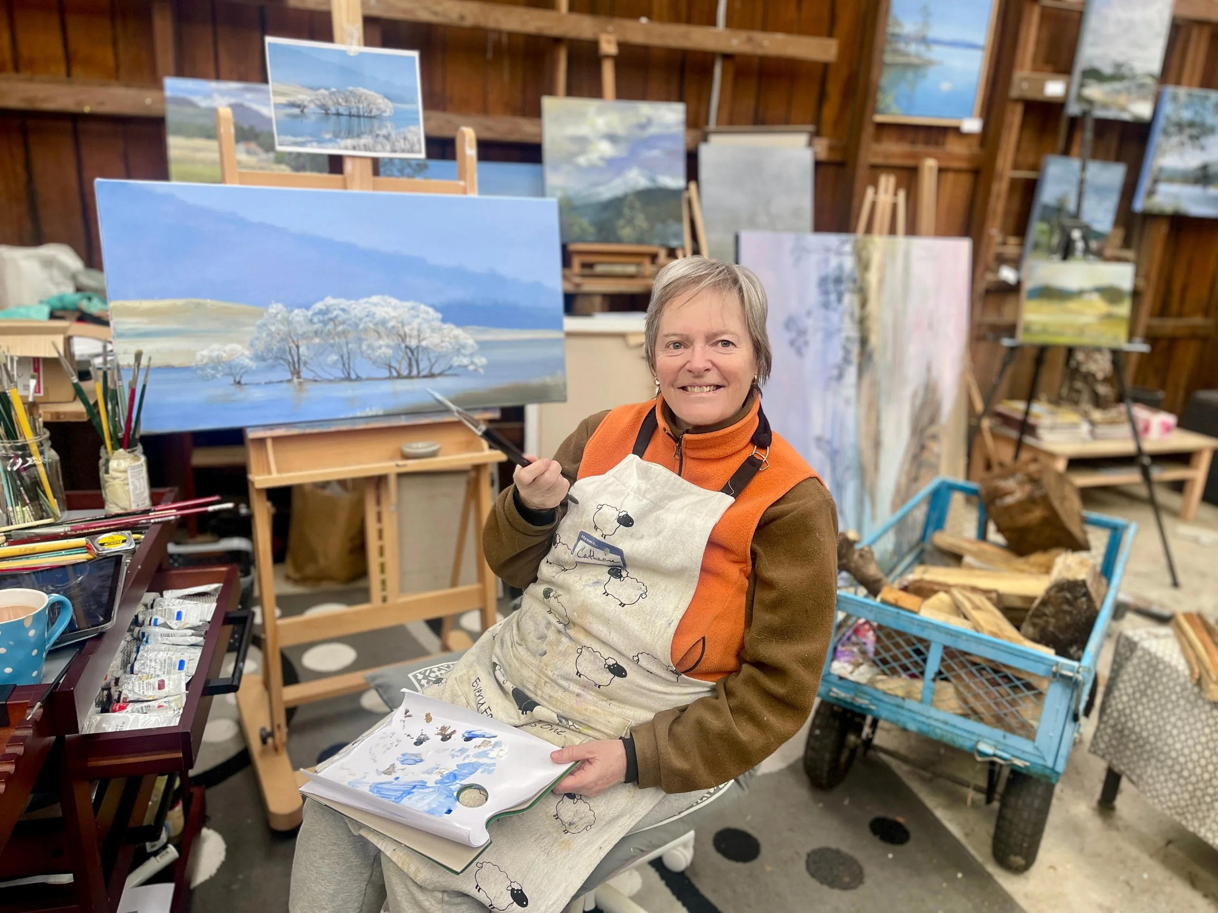 An older woman with short gray hair smiling while holding an open watercolor art book, standing in an art studio with wooden walls. She wears an orange jacket and a beige apron with black drawings, surrounded by easels, canvases, and art supplies, including tubes of paint and brushes. There are several landscape paintings of trees, water, and mountains displayed on the walls and easels.