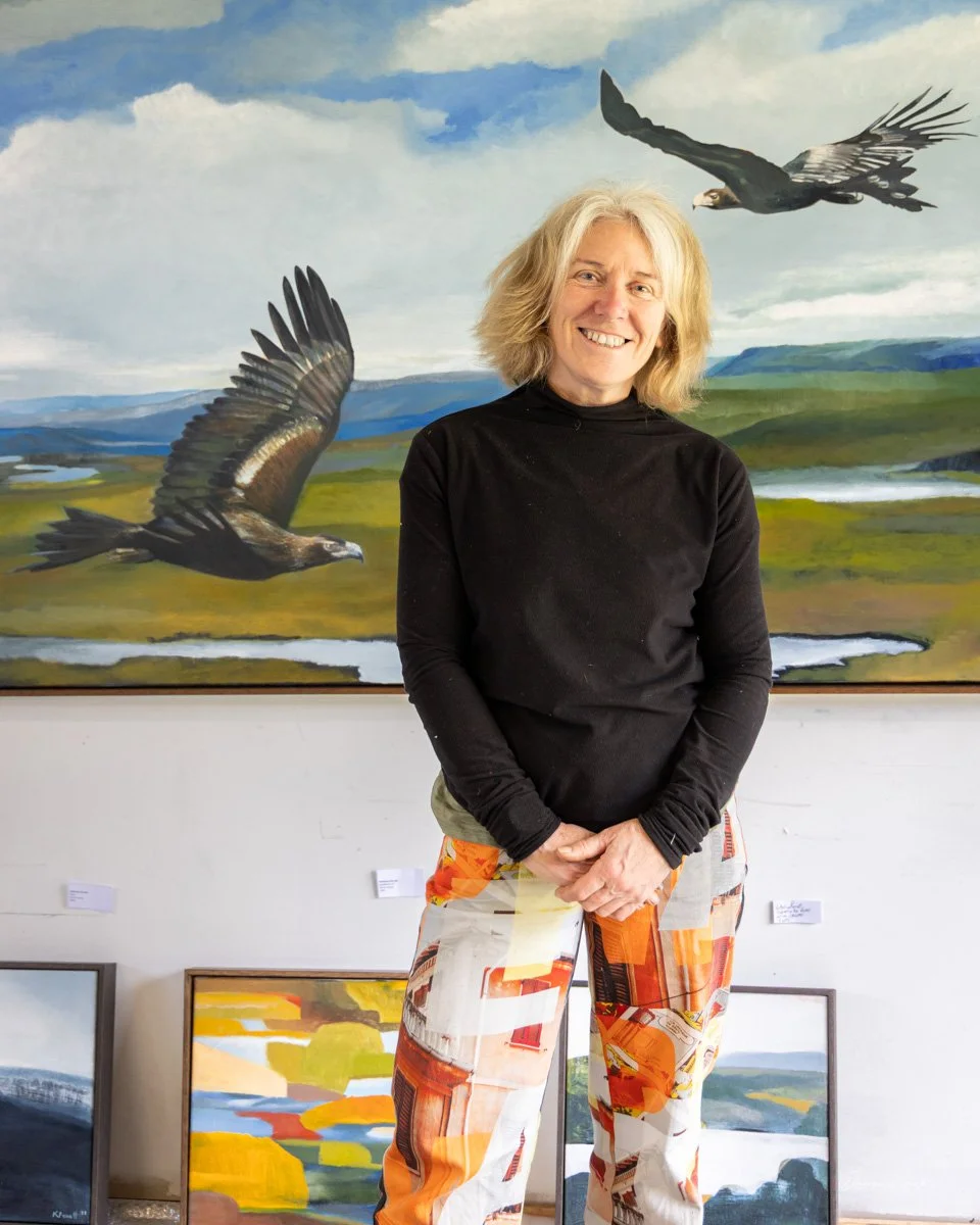 A woman with blonde hair wearing a black long-sleeve top and colorful patterned pants, standing in front of a large landscape painting featuring mountains, water, and flying birds, including two prominent eagles.