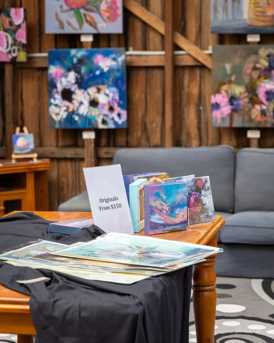 Table displaying art prints and books with a sign indicating 'Originals From $150' in an art gallery with colorful paintings on wooden walls.