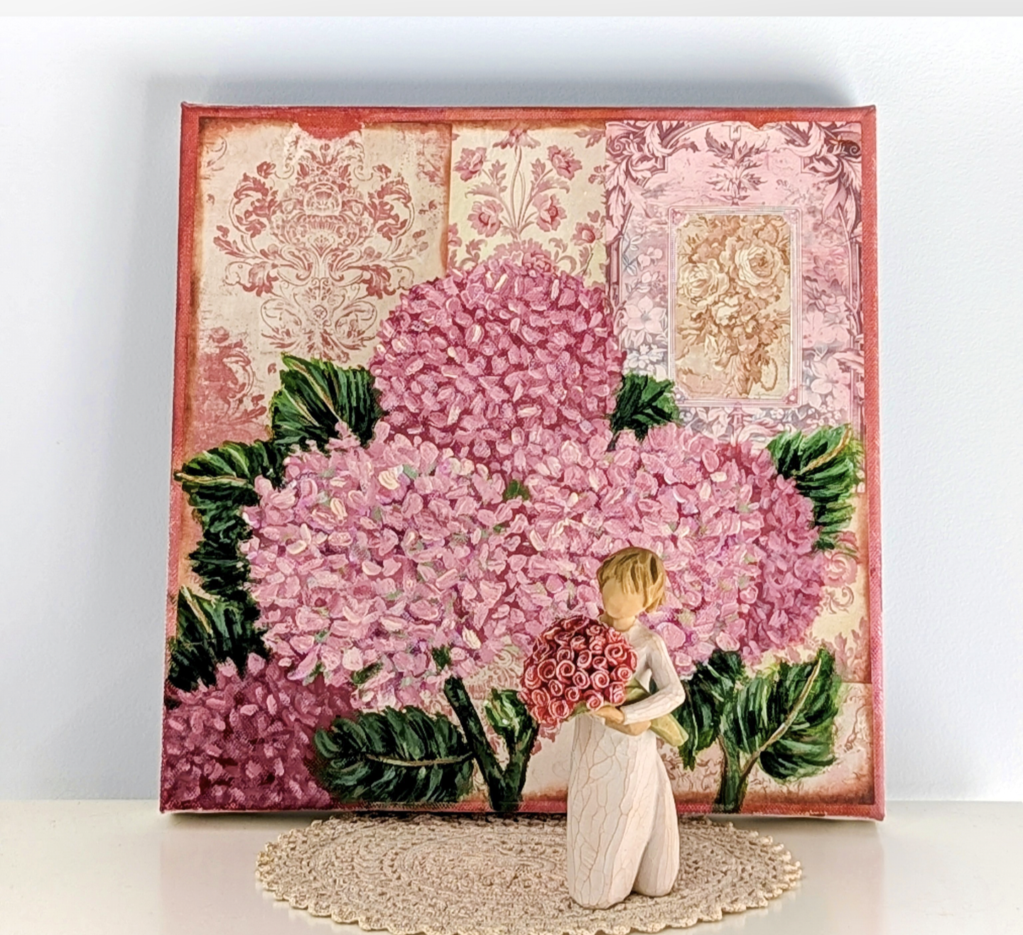 A decorative artwork featuring a small figurine holding a bouquet of pink roses, set against a background of pink-themed floral wallpaper and illustrations, with a lace doily underneath.