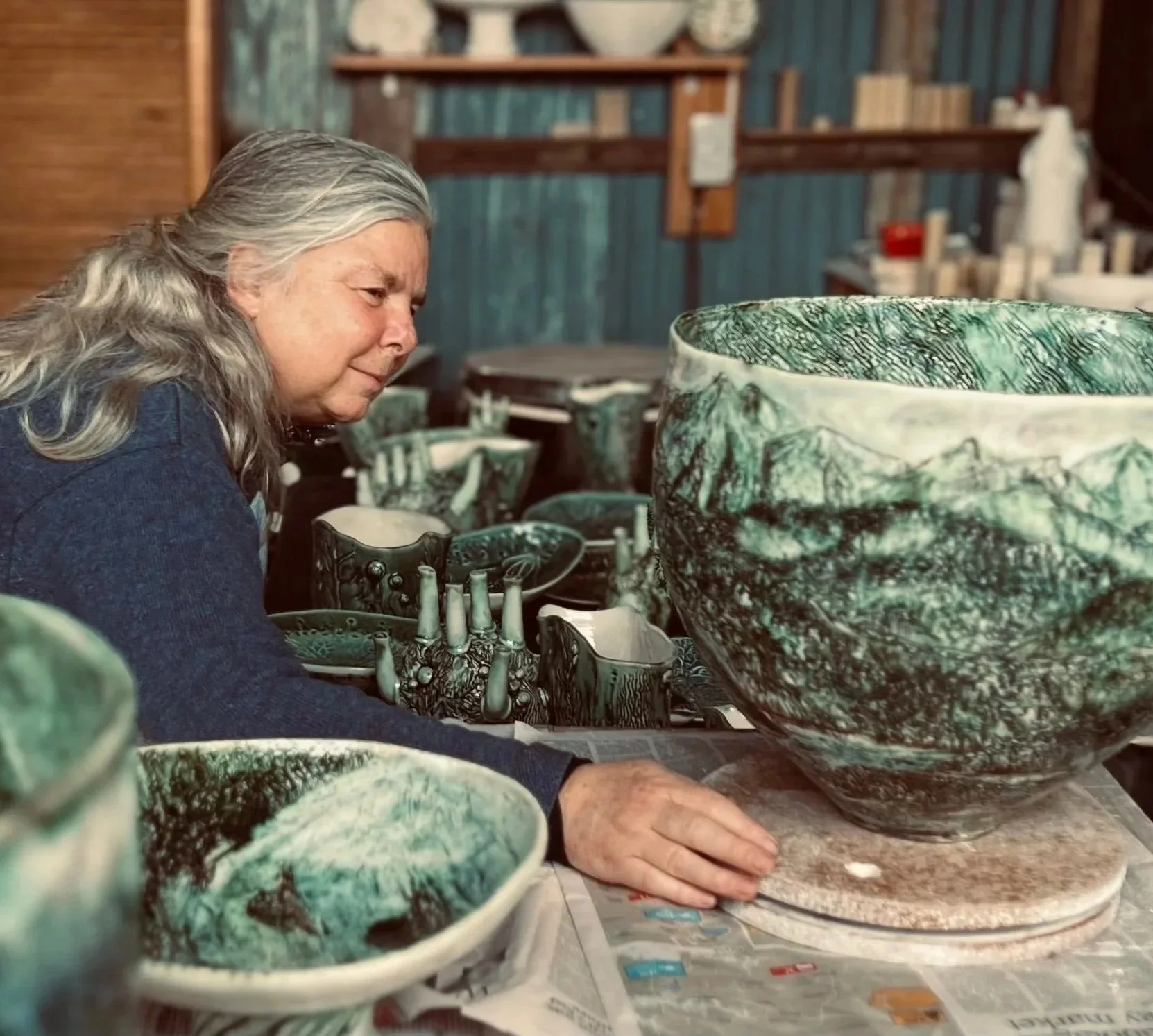 An elderly woman with gray hair, wearing a dark blue sweater, working on a large, green marbled ceramic bowl in a workshop. Several other smaller similar bowls are on the table nearby, with shelves and tools in the background.