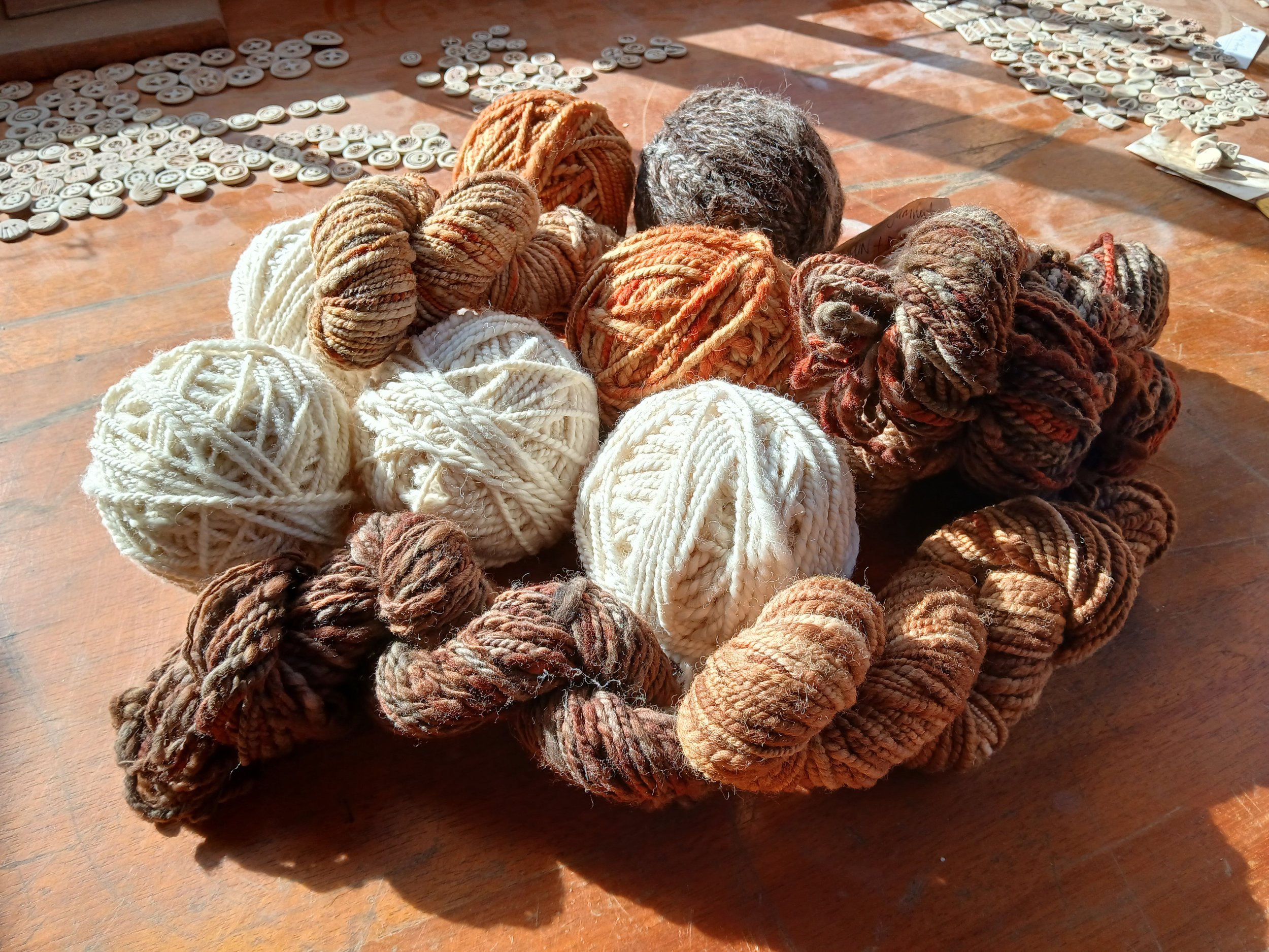 A pile of yarn balls in various shades of white, beige, orange, and brown, placed on a wooden surface with scattered buttons and sewing supplies in the background.