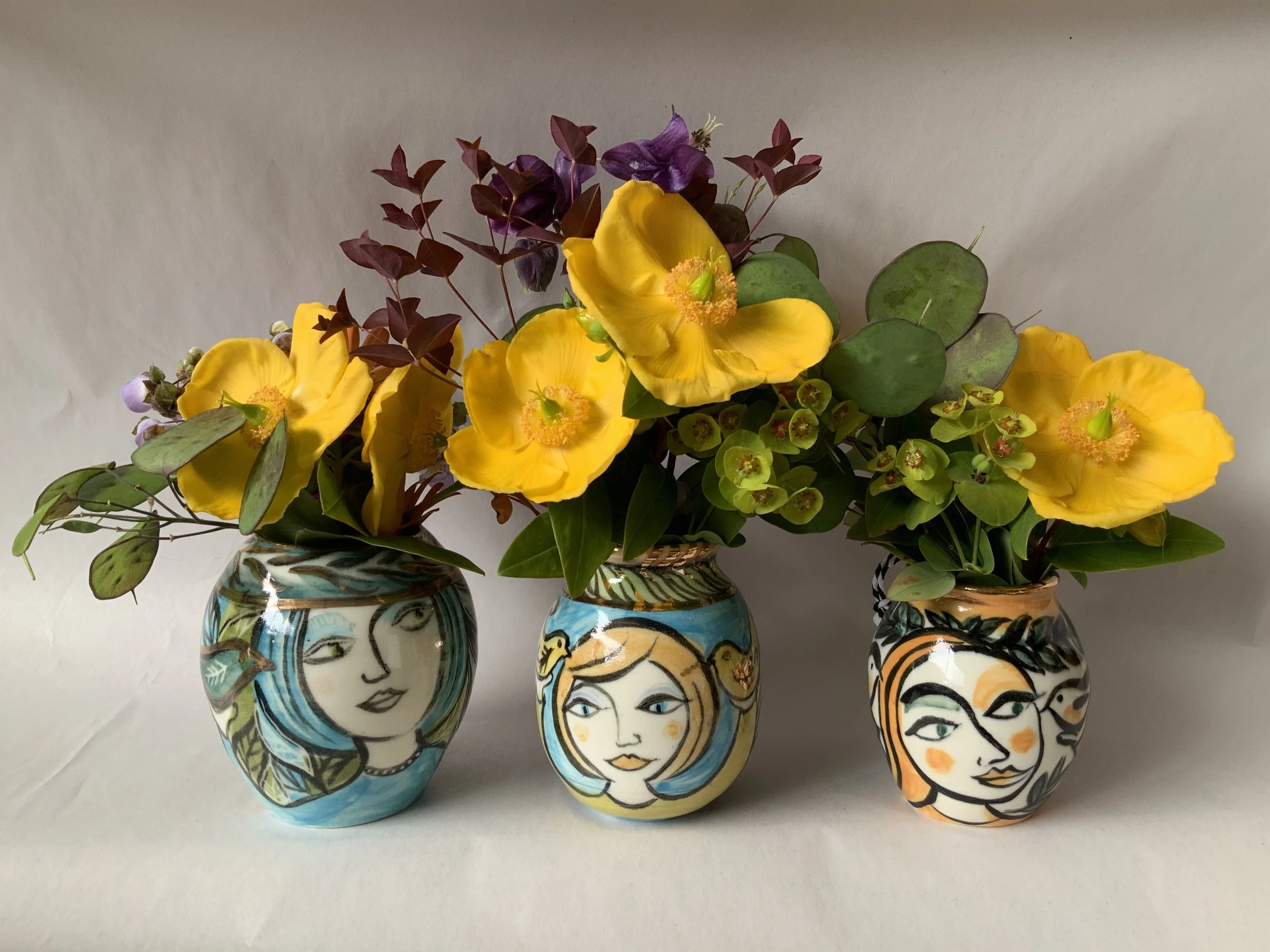Three decorative vases with painted faces design, holding bright yellow flowers and purple and green foliage, arranged against a plain background.