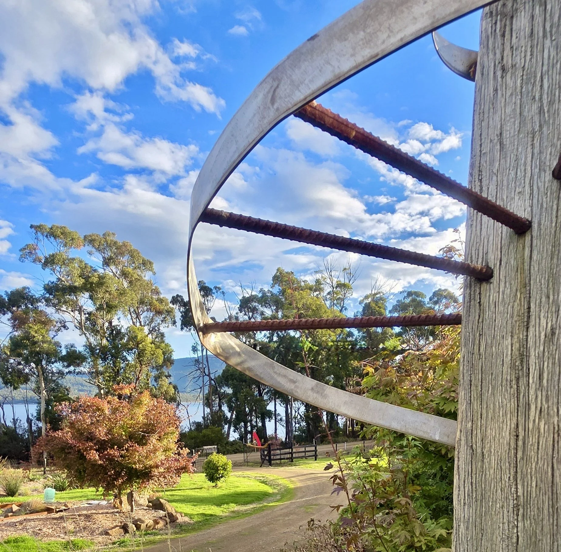 Close-up of a wooden post with metal rings attached, set against a background of trees, grass, a bench, and a cloudy blue sky.