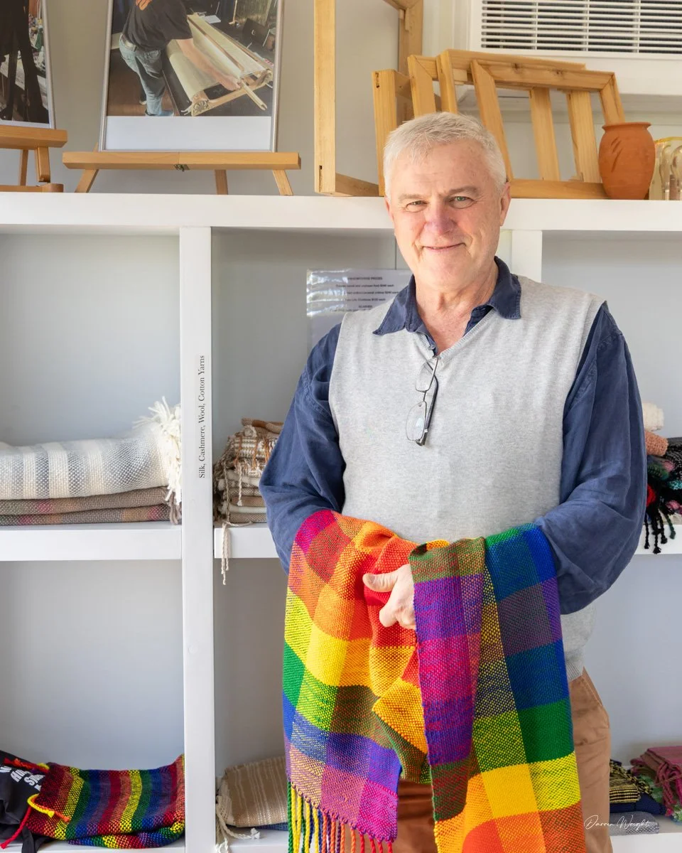 A man holding a colorful plaid textile in a store with displayed woven fabrics and artwork in the background.