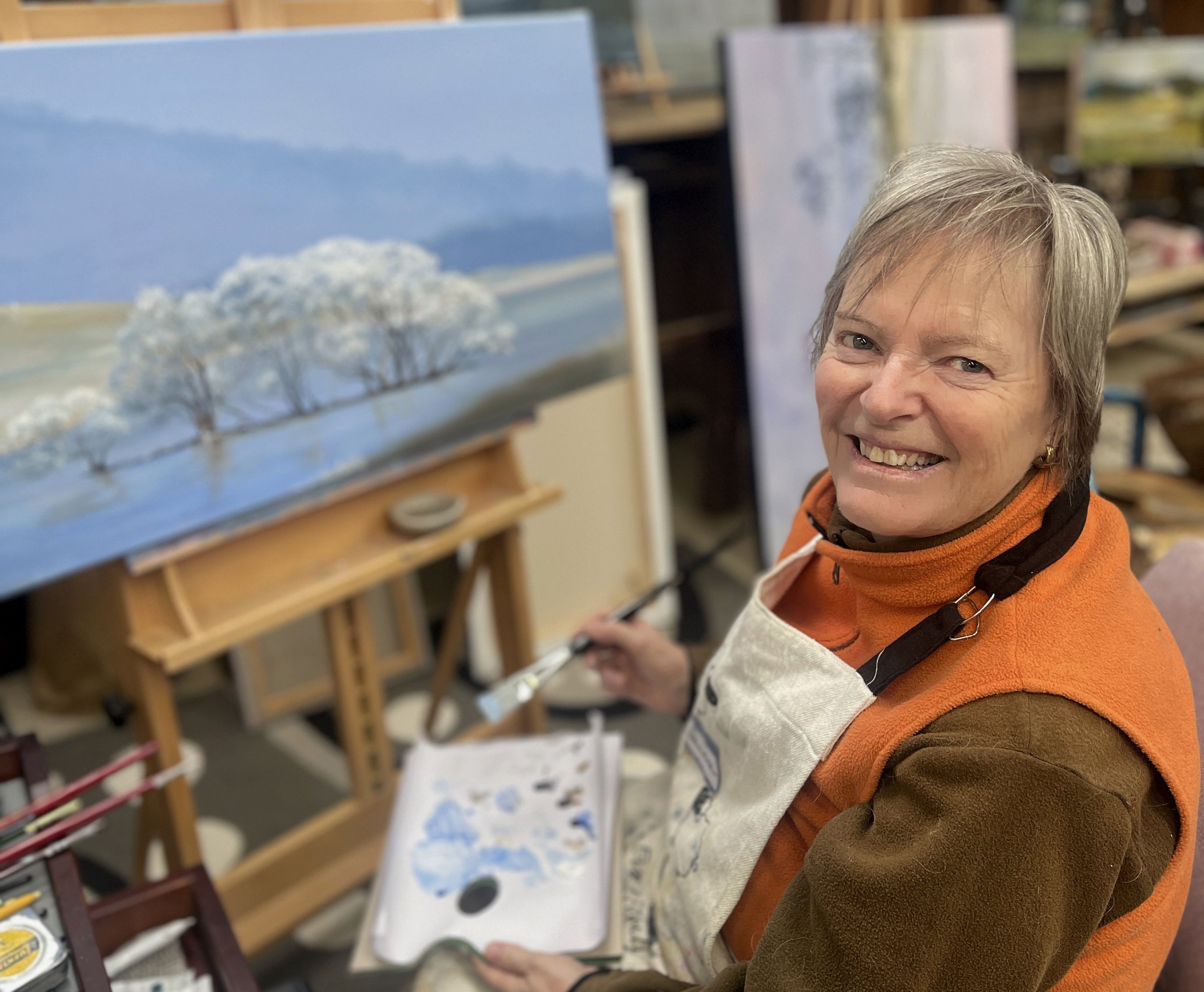 A smiling woman with short gray hair wearing an orange vest and apron, painting a landscape scene of trees under a blue sky on a canvas in an art studio.