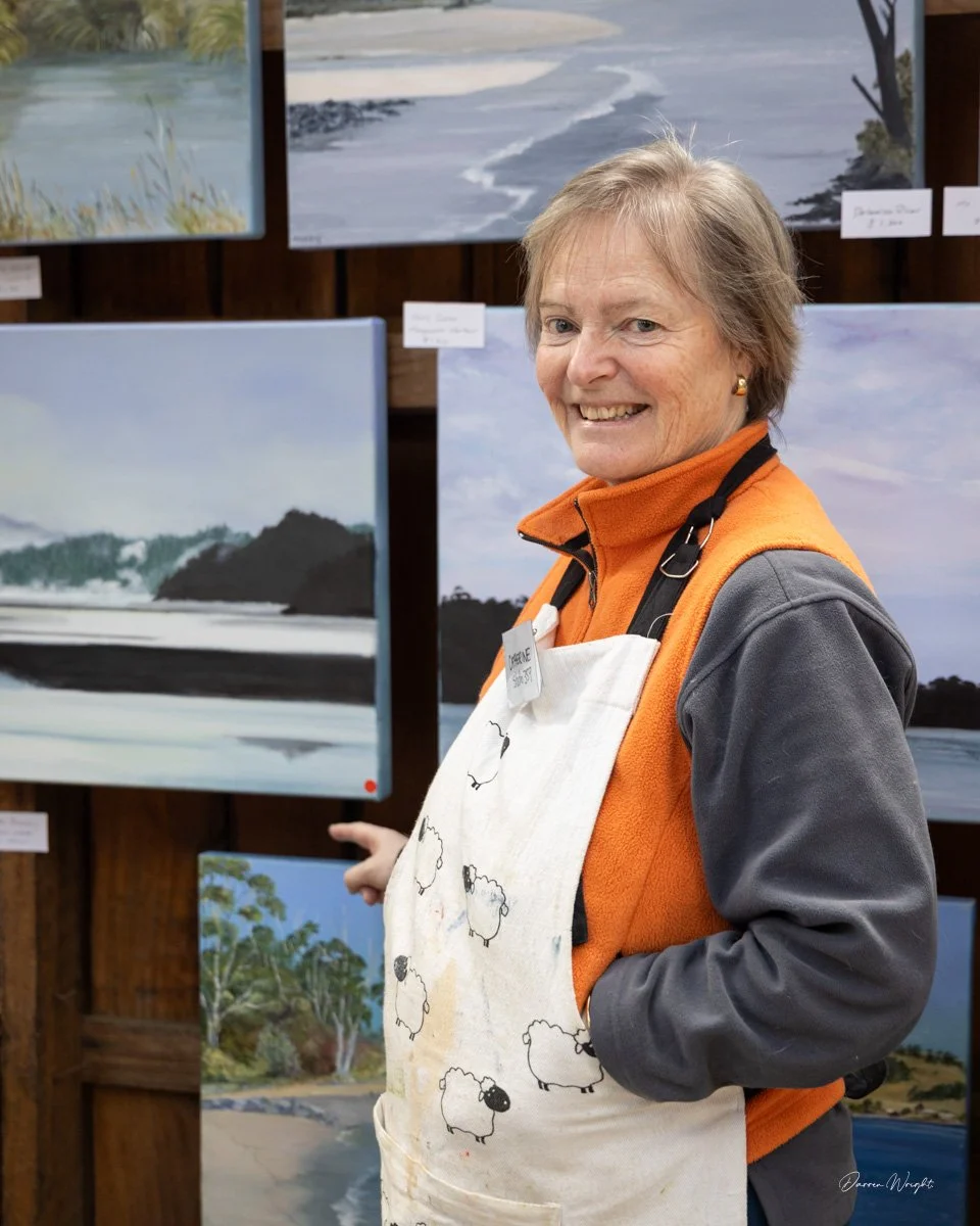 An elderly woman with short gray hair is smiling and standing in front of paintings of nature scenes at an art show, wearing an orange and gray jacket and an apron with sheep patterns.