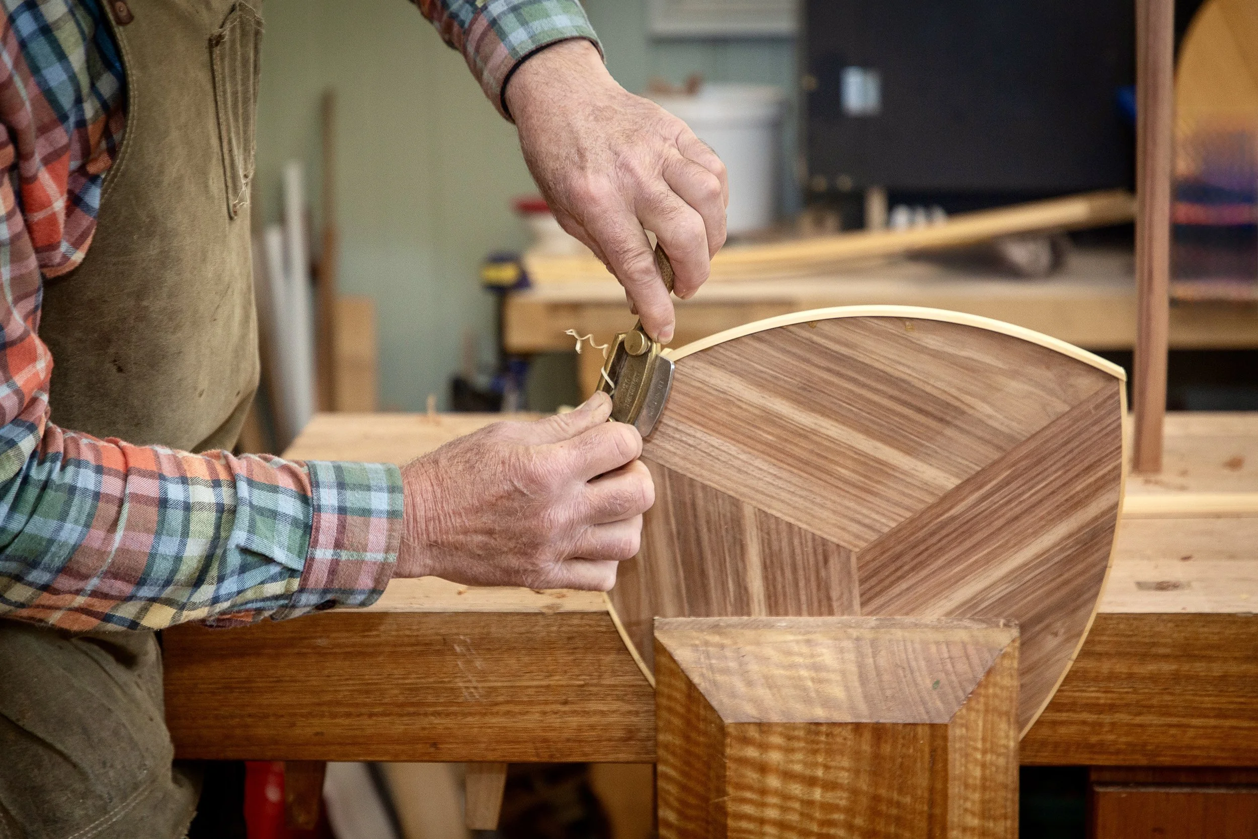 A person using a hand tool on a wooden furniture piece in a woodworking workshop.
