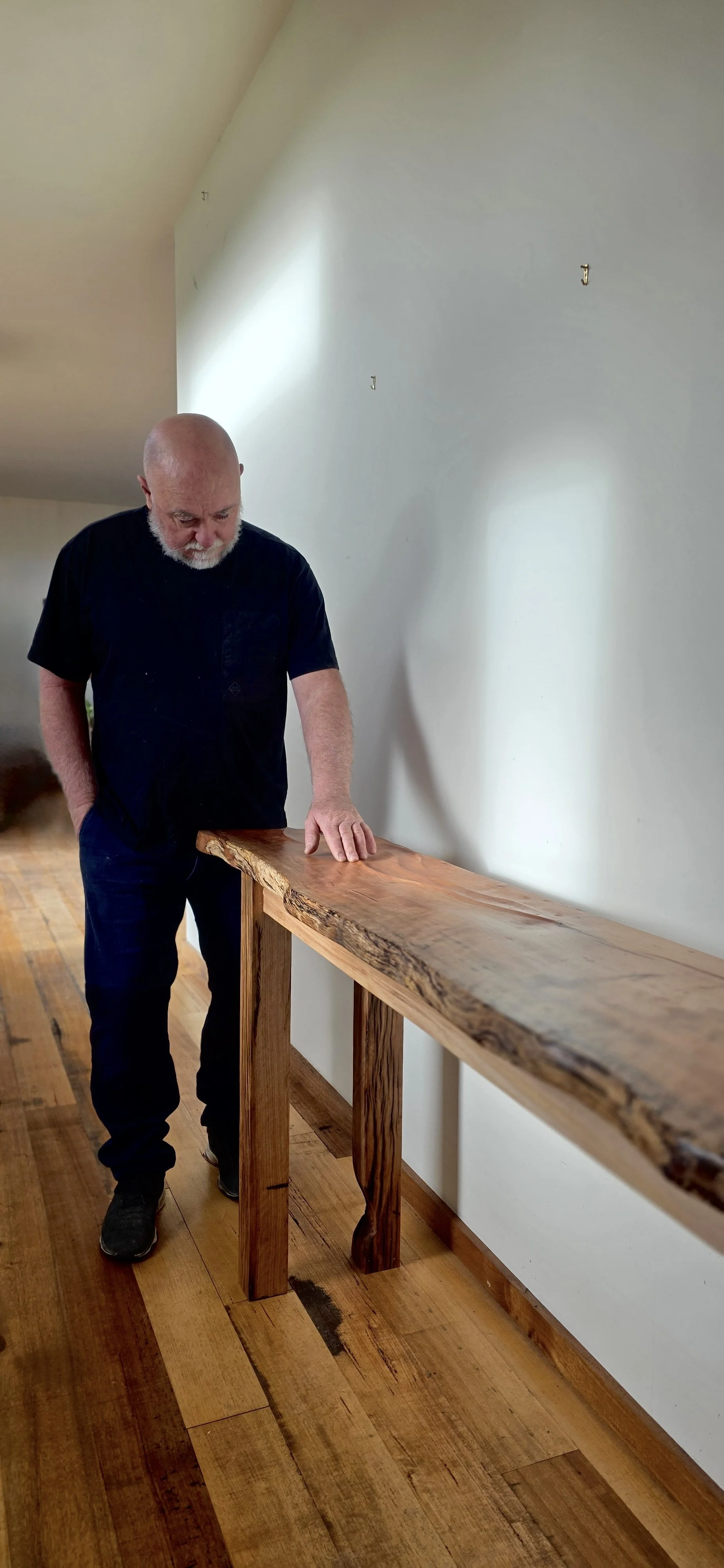 An older man with a bald head and white beard standing next to a wooden hand railing, looking down at the railing with his right hand resting on it and left hand in his pocket in a room with wood flooring and a white wall.