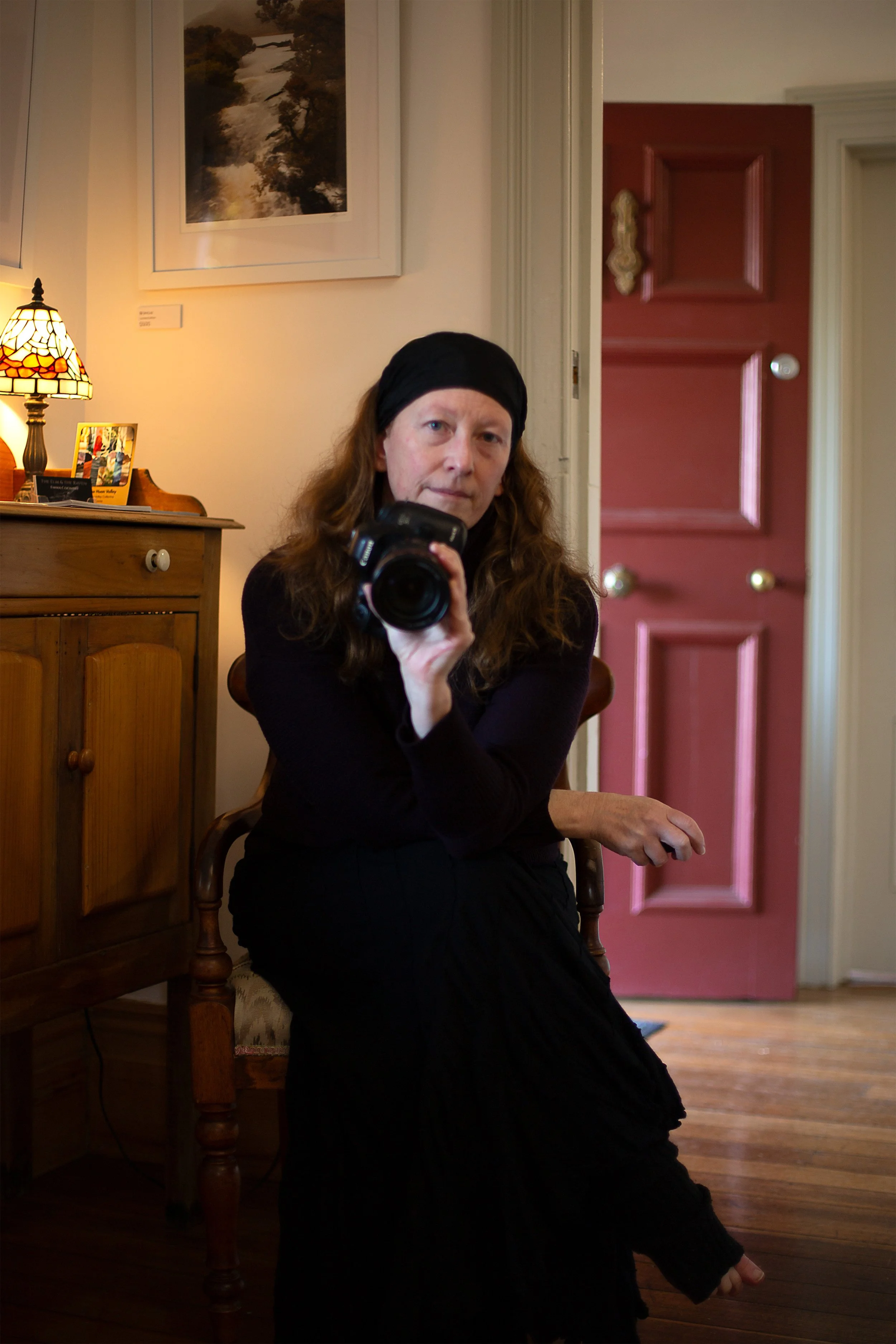 A woman with long brown hair, wearing a black headscarf and black clothing, is sitting inside a house and looking into a mirror while holding a camera.
