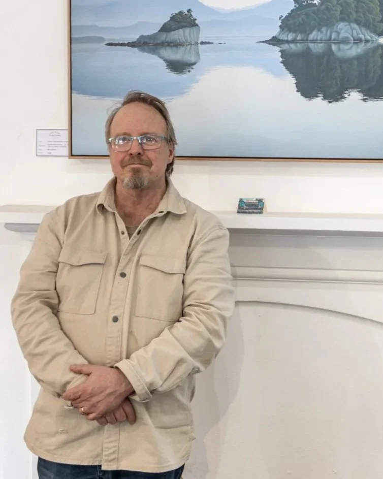 A man with glasses and a beige jacket standing in front of a white wall with a large framed painting of a peaceful lakeside scene with islands and trees.